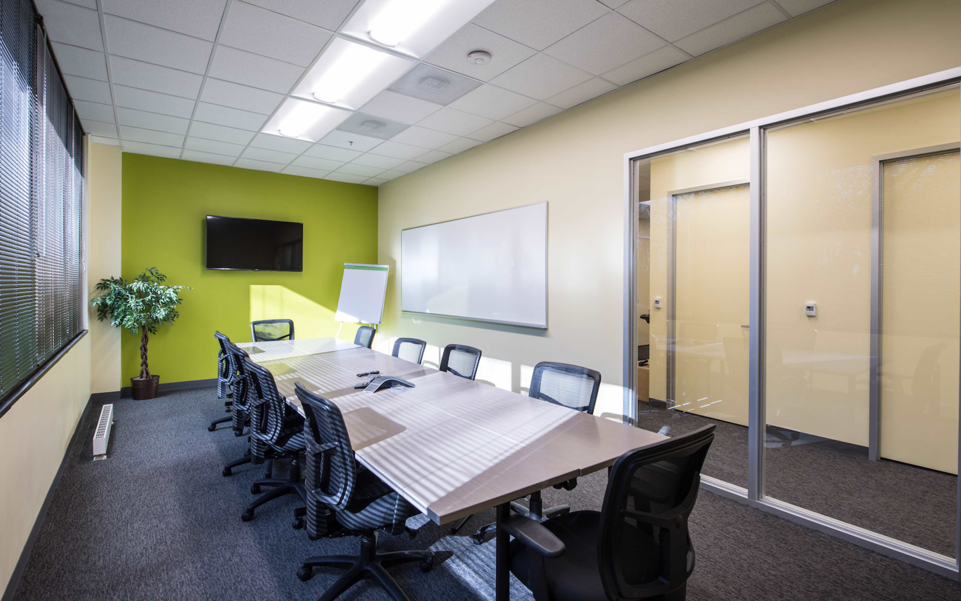 The image shows a modern conference room with a long table surrounded by office chairs, a whiteboard, a television screen on the wall, and a glass-walled meeting room adjacent to it.