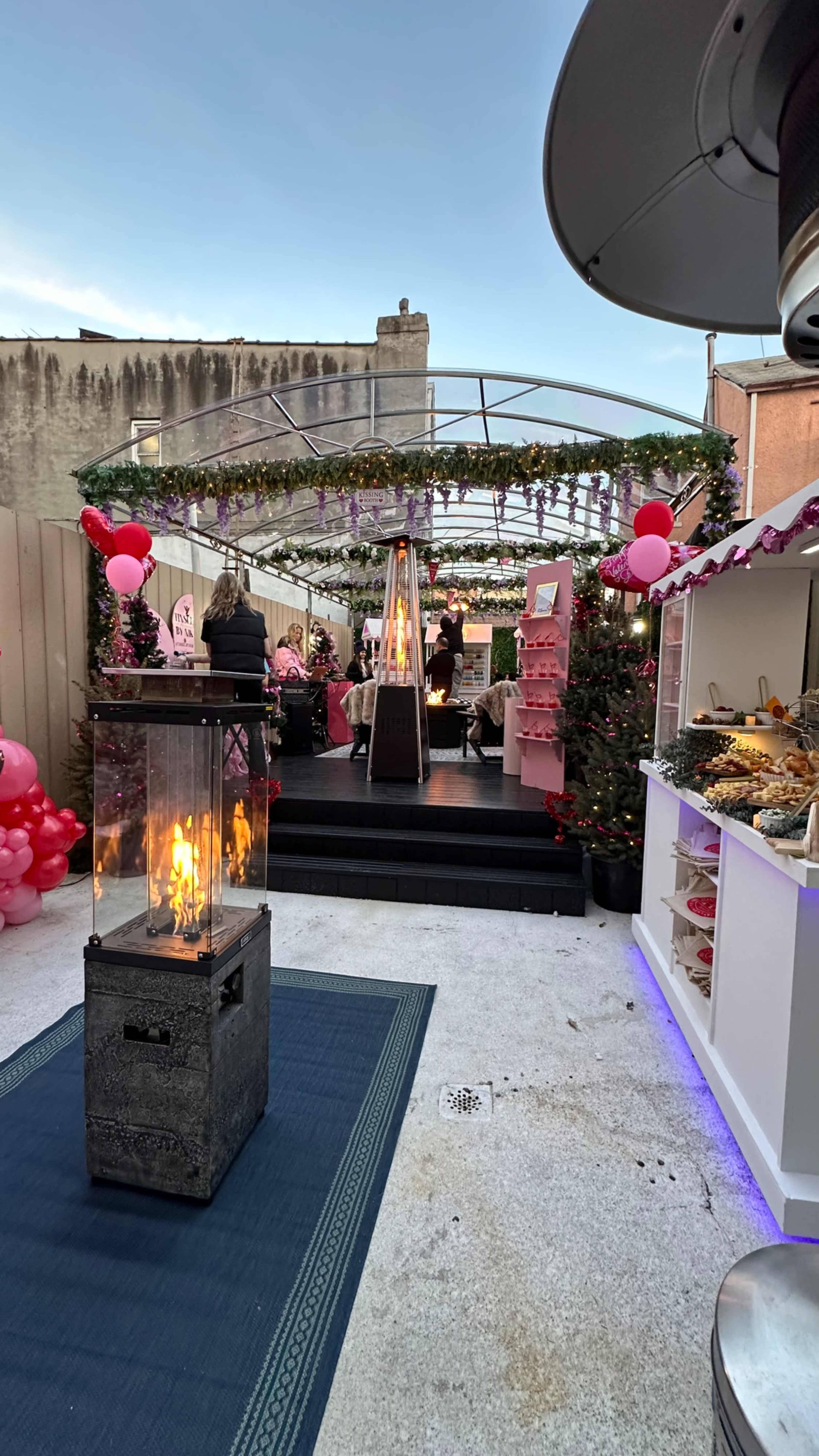 The scene depicts a decorated outdoor event space with festive pink balloons, a heating unit, and a buffet table adorned with food, surrounded by plants and a canopy of greenery.