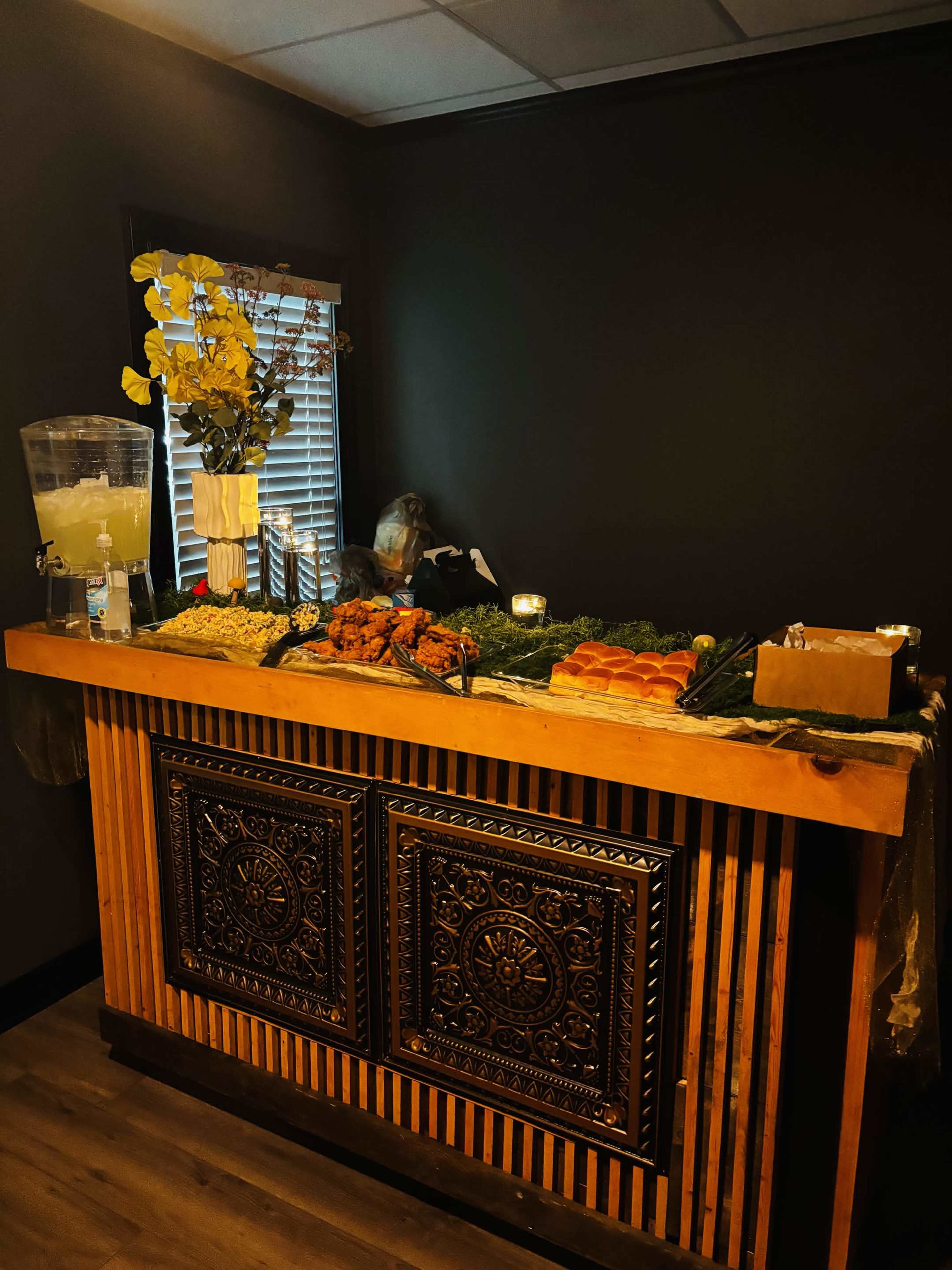 The image depicts a wooden service bar adorned with a variety of food items, including fruits and snacks, along with a large beverage dispenser, surrounded by decorative elements.