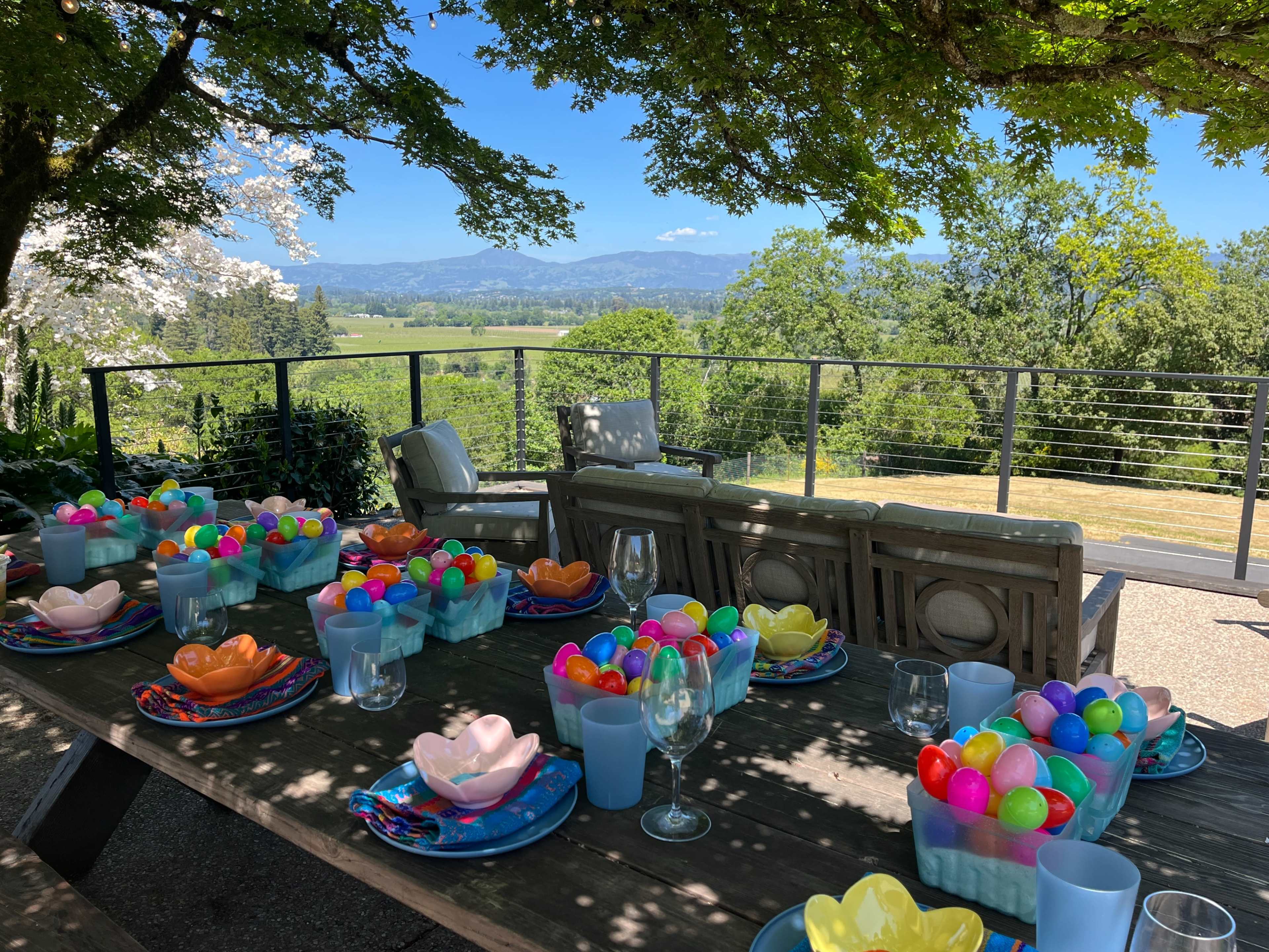 A long outdoor dining table is set with colorful bowls filled with plastic eggs, overlooking a scenic view of rolling hills and trees under a clear blue sky.