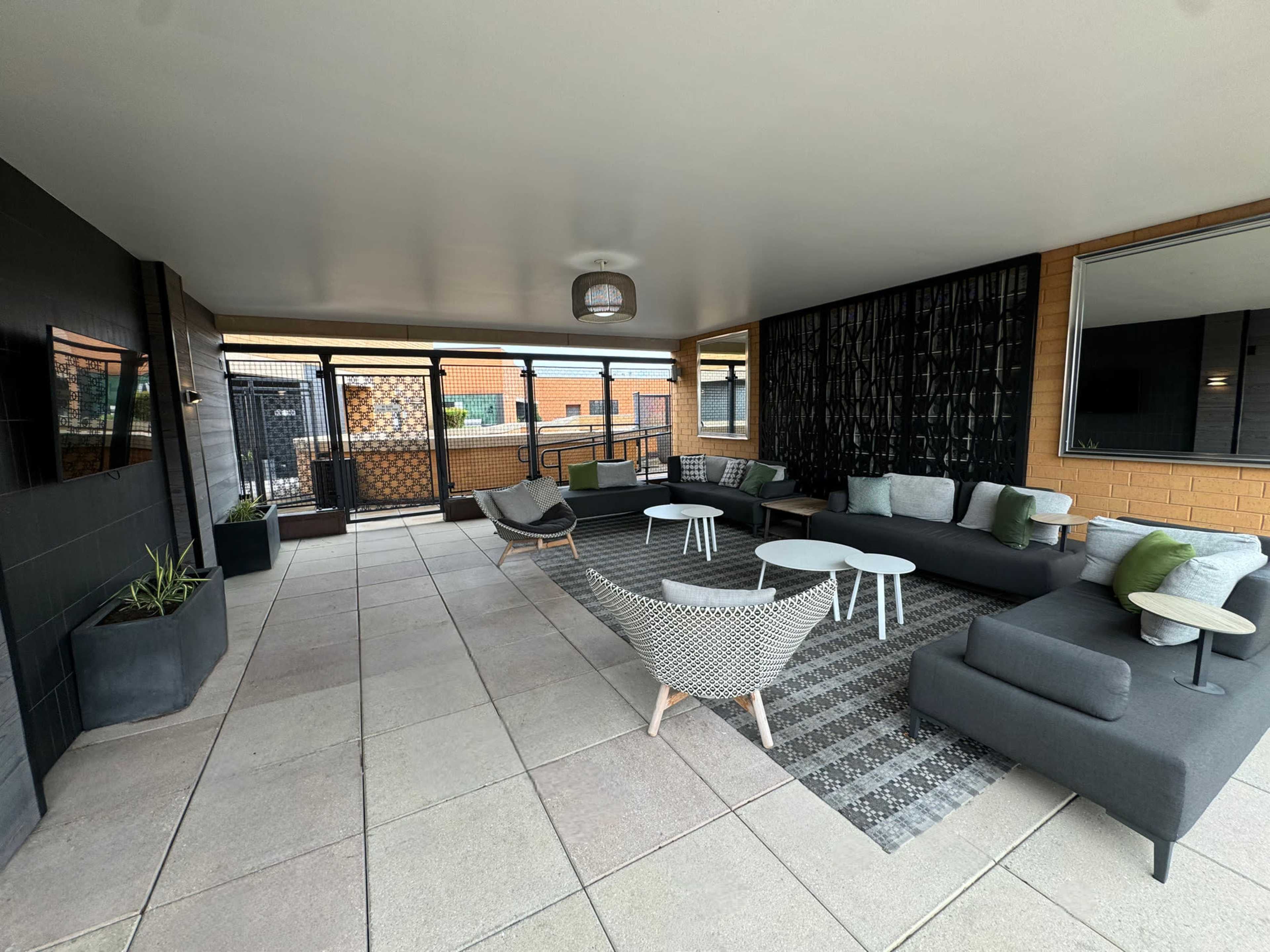 A modern outdoor lounge area with seating arrangements, a patterned rug, and green accents against a neutral backdrop.