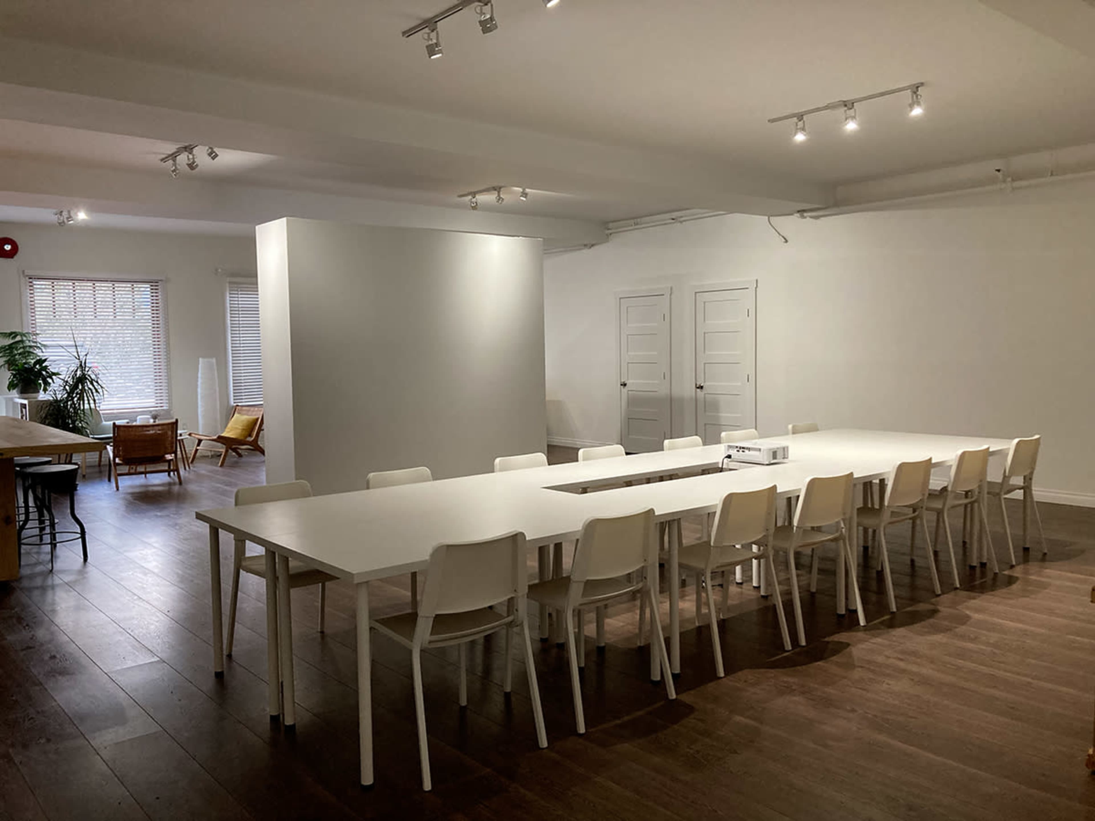 A long, white table with multiple chairs surrounds it in a well-lit, minimalist room featuring a separate partition and wooden flooring.
