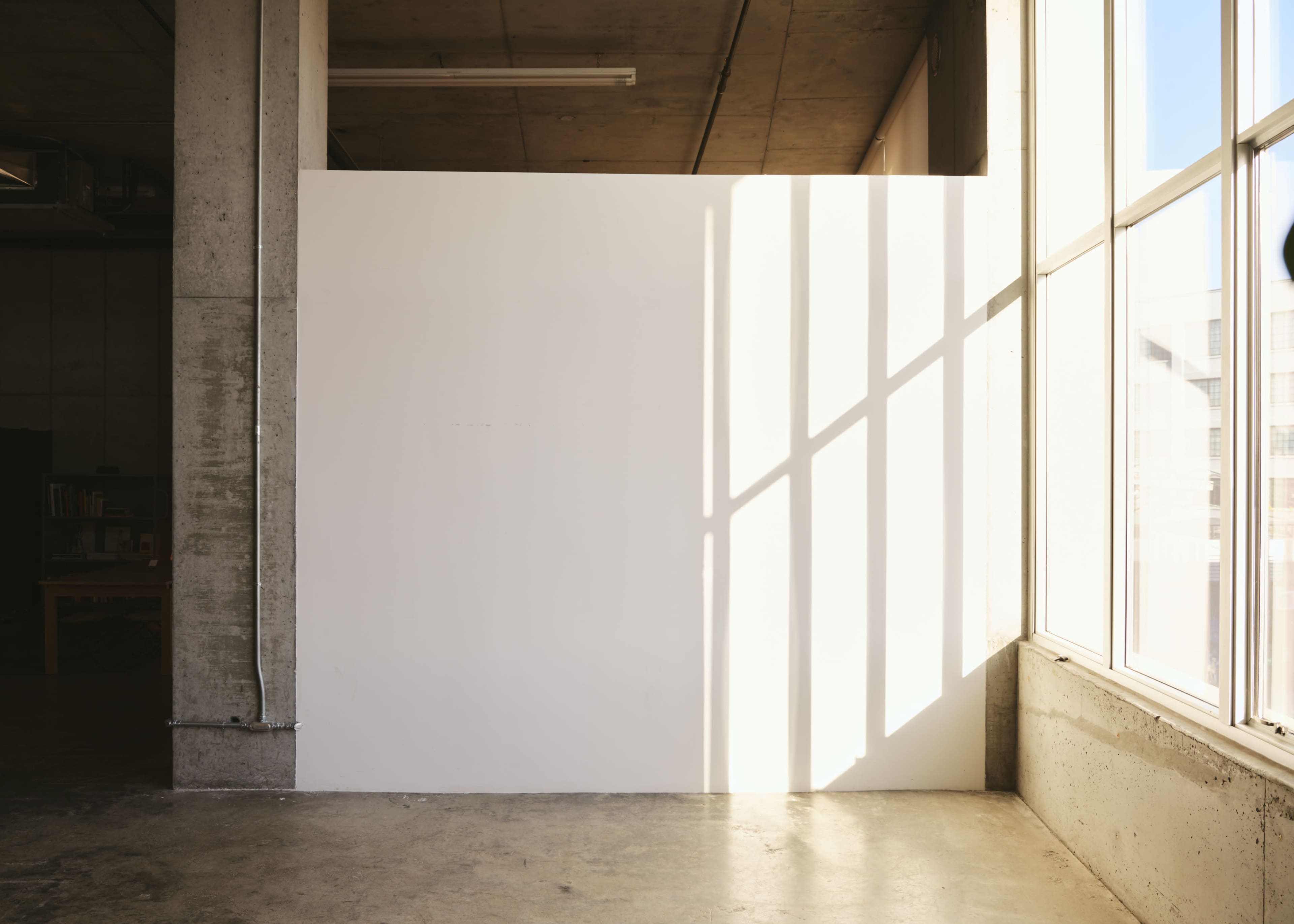 The image shows a white wall illuminated by sunlight streaming through large windows, with concrete elements in the surrounding space.
