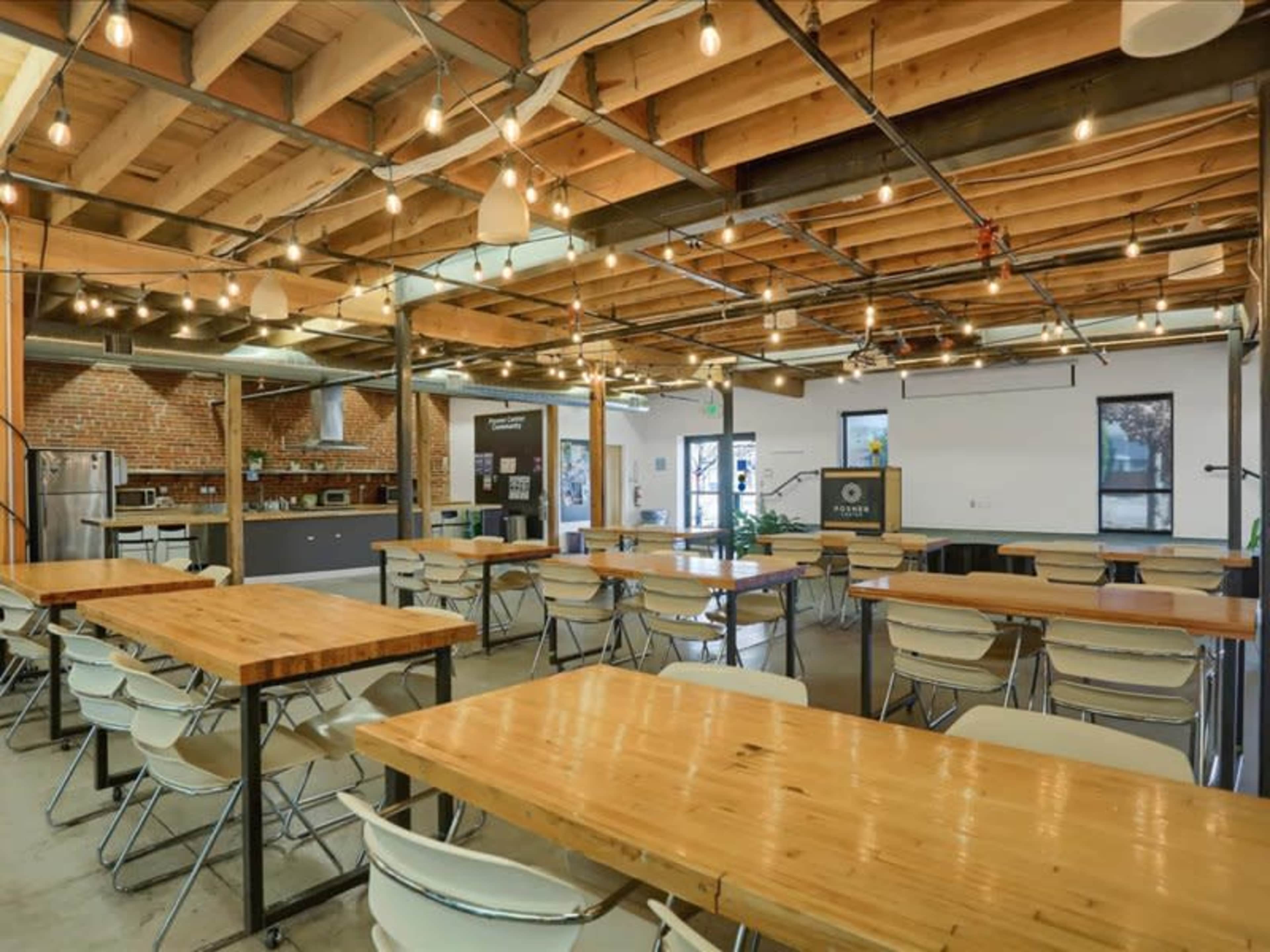 The image shows a spacious, modern workspace with wooden tables and chairs, industrial lighting, and exposed brick walls.