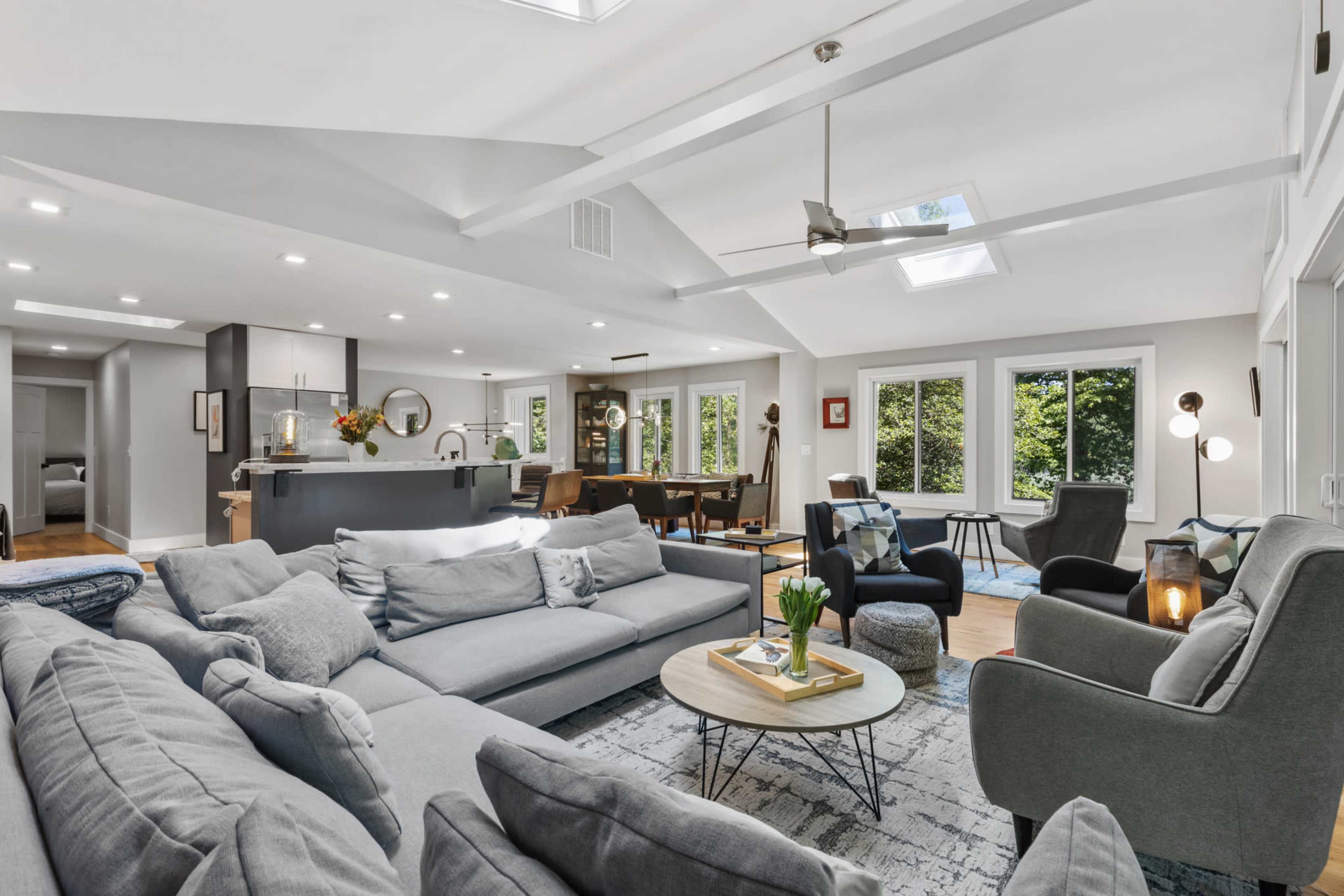 The image shows a modern, open-concept living area with a large sectional sofa, a round coffee table, and kitchen space in the background, illuminated by natural light from skylights and windows.