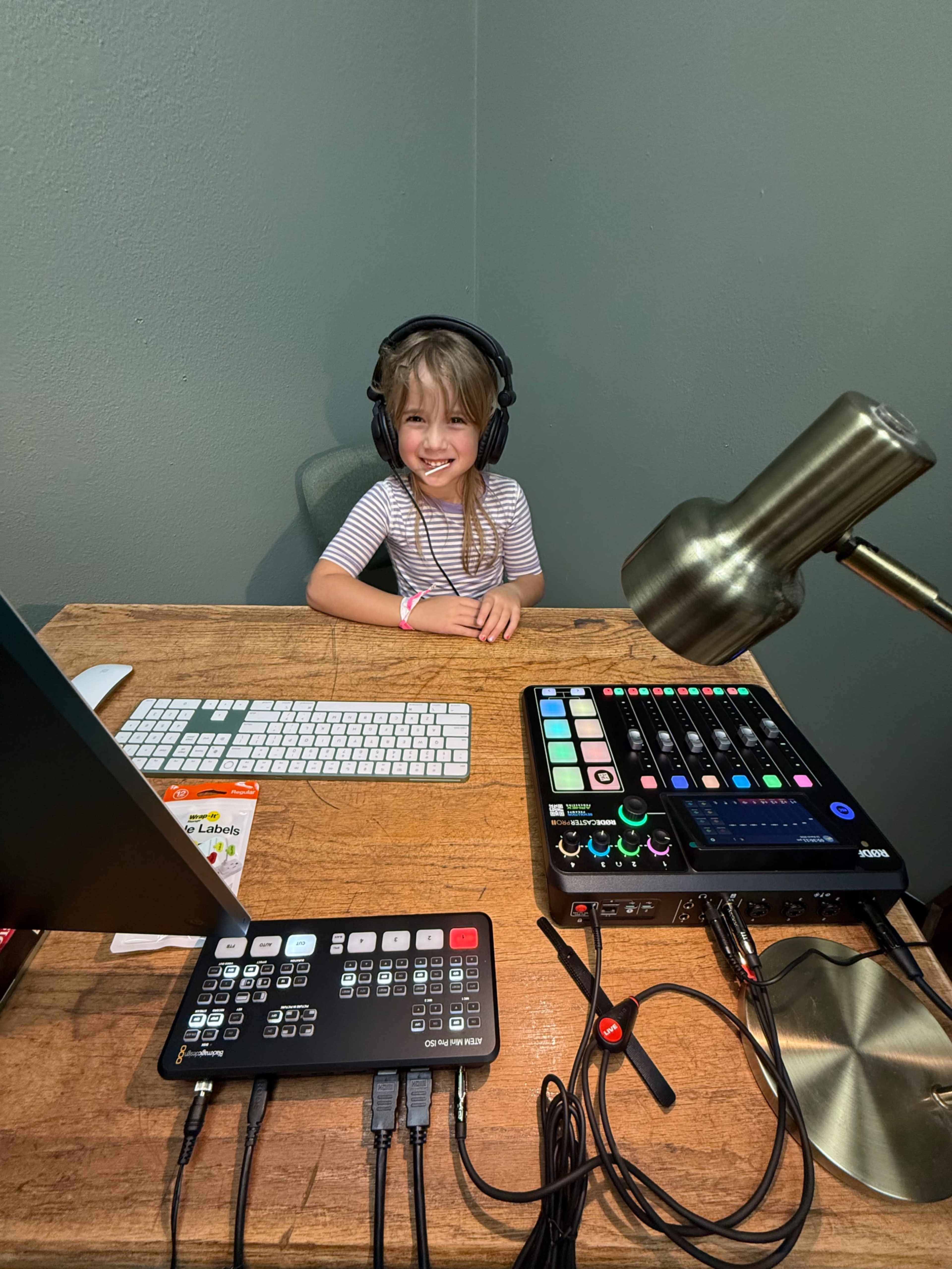 A child wearing headphones sits at a wooden desk with a computer, audio equipment, and a desk lamp.
