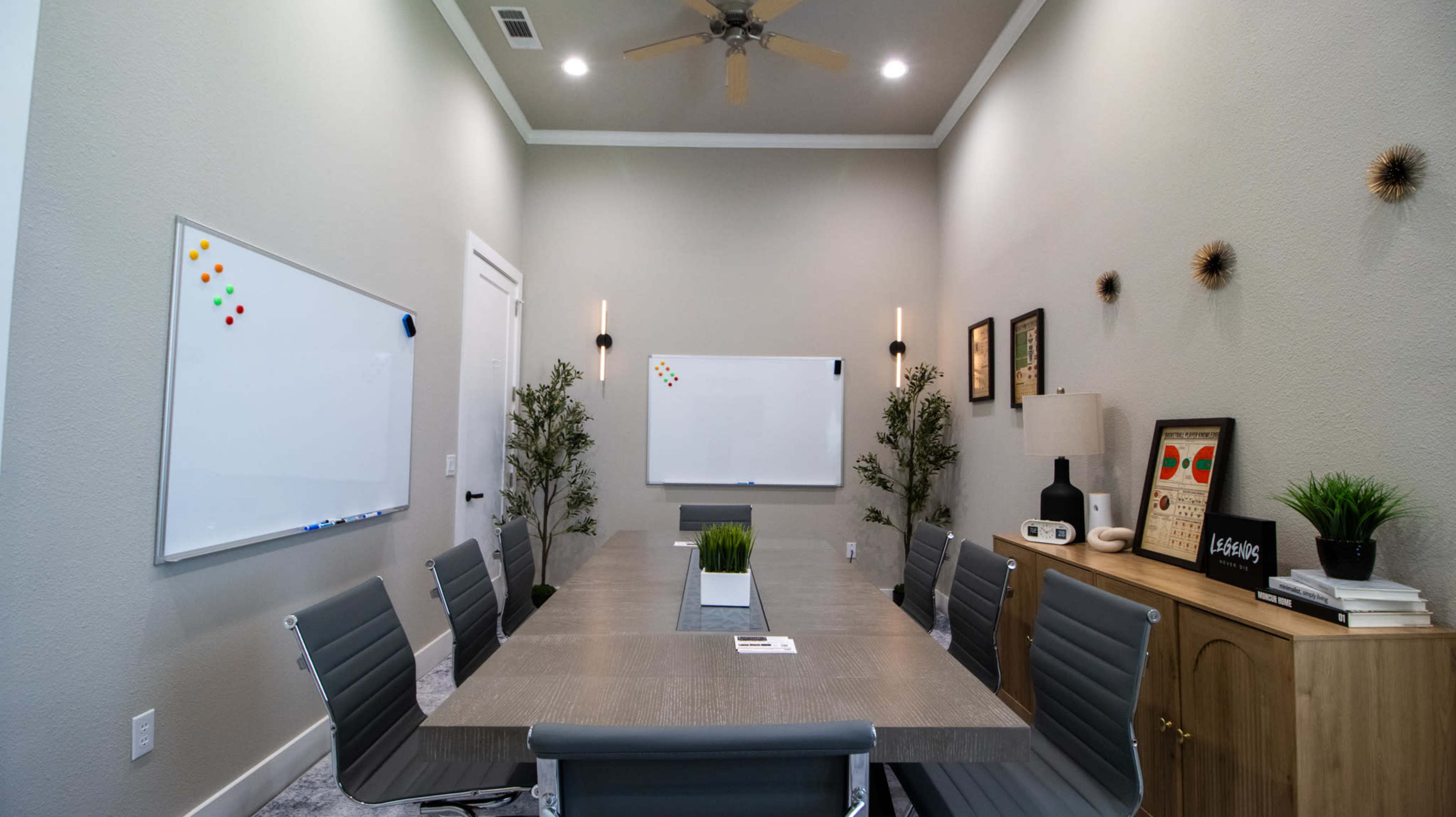 A modern conference room features a long wooden table surrounded by gray chairs, with whiteboards on the walls and decorative plants in the corners.