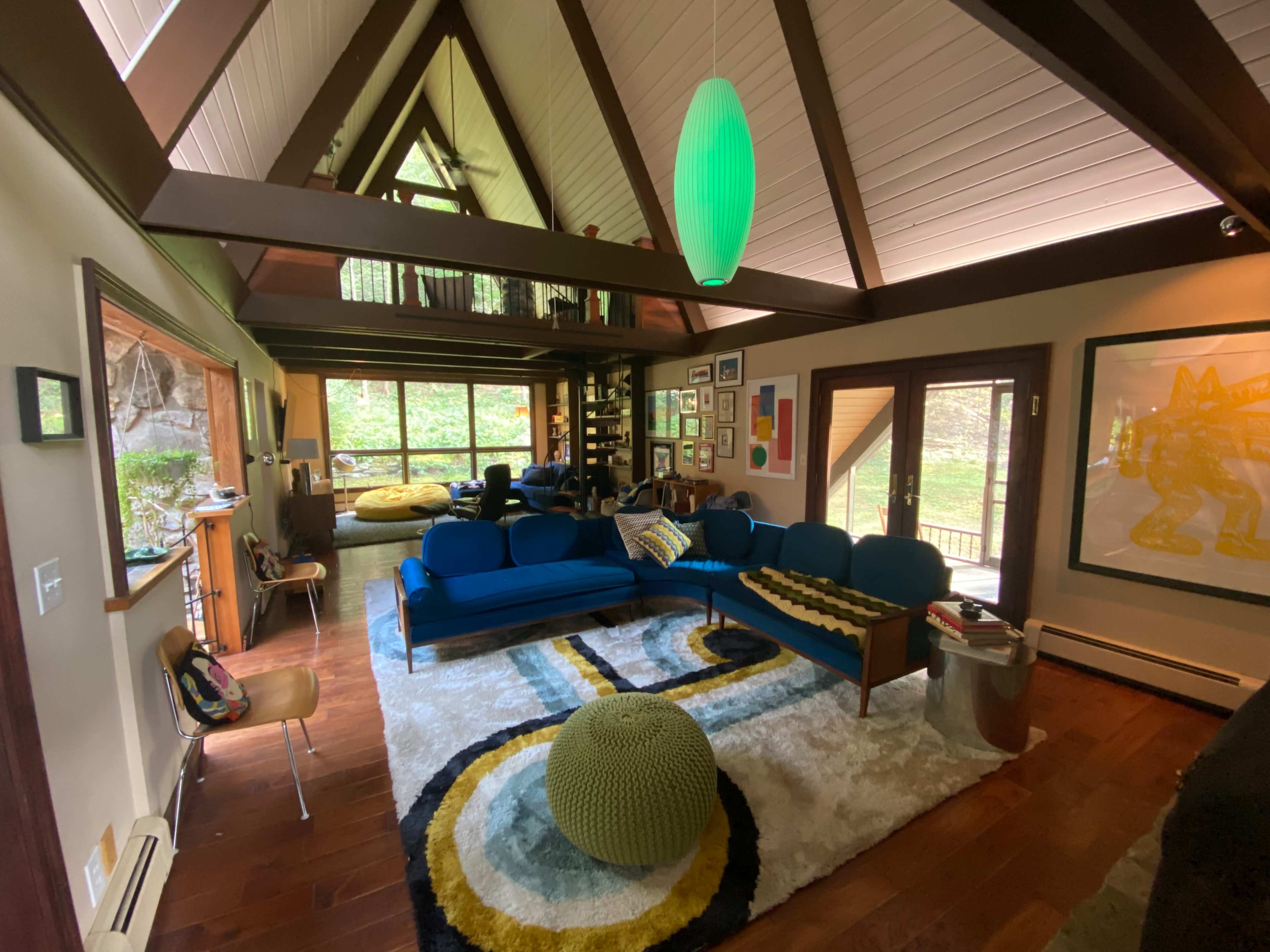 A cozy living room with a triangular wooden ceiling, blue sofas, a patterned rug, and large windows providing views of a green outdoor space.
