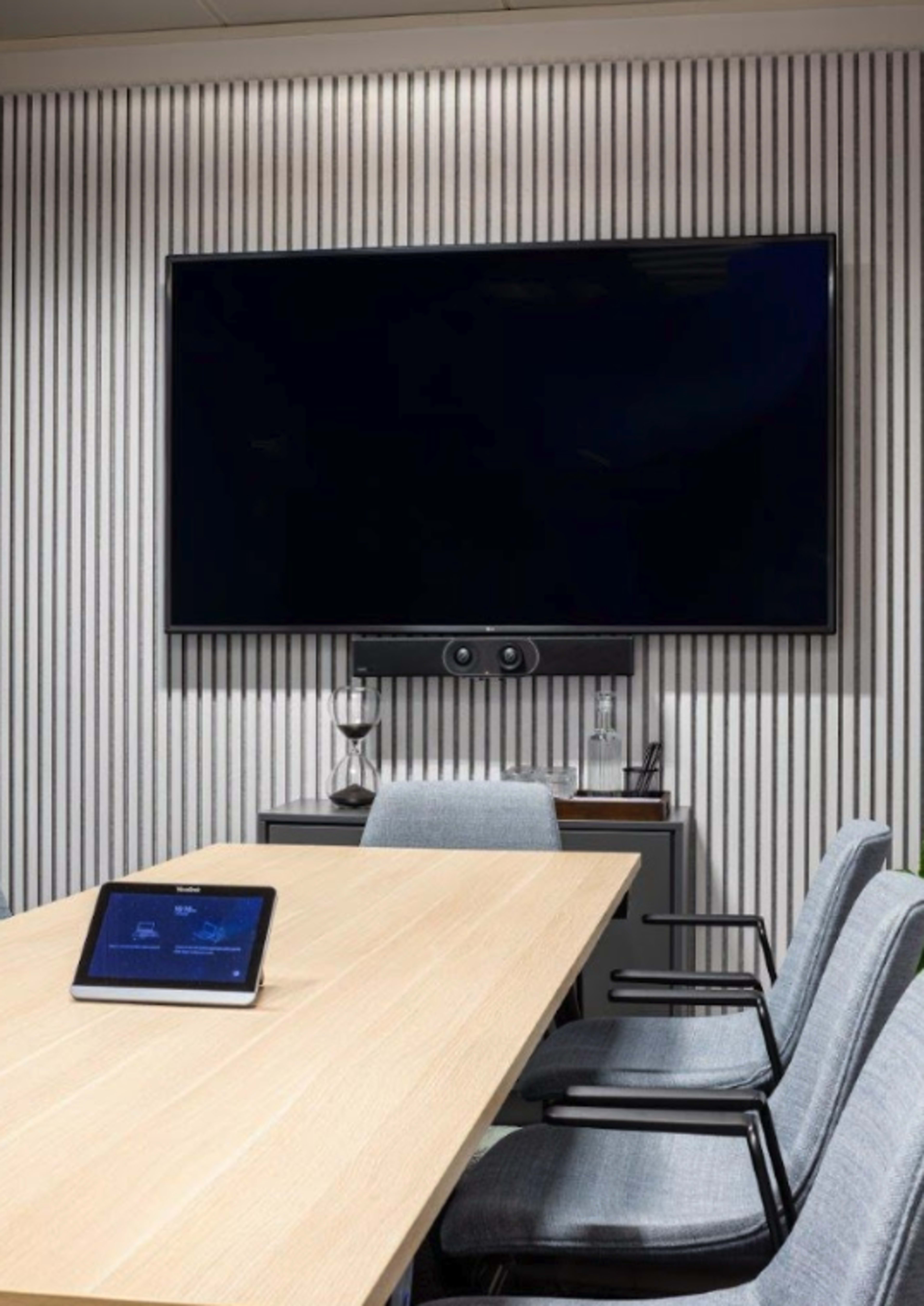 A modern conference room features a wooden table, several gray chairs, a large wall-mounted television, and a tablet placed on the table.