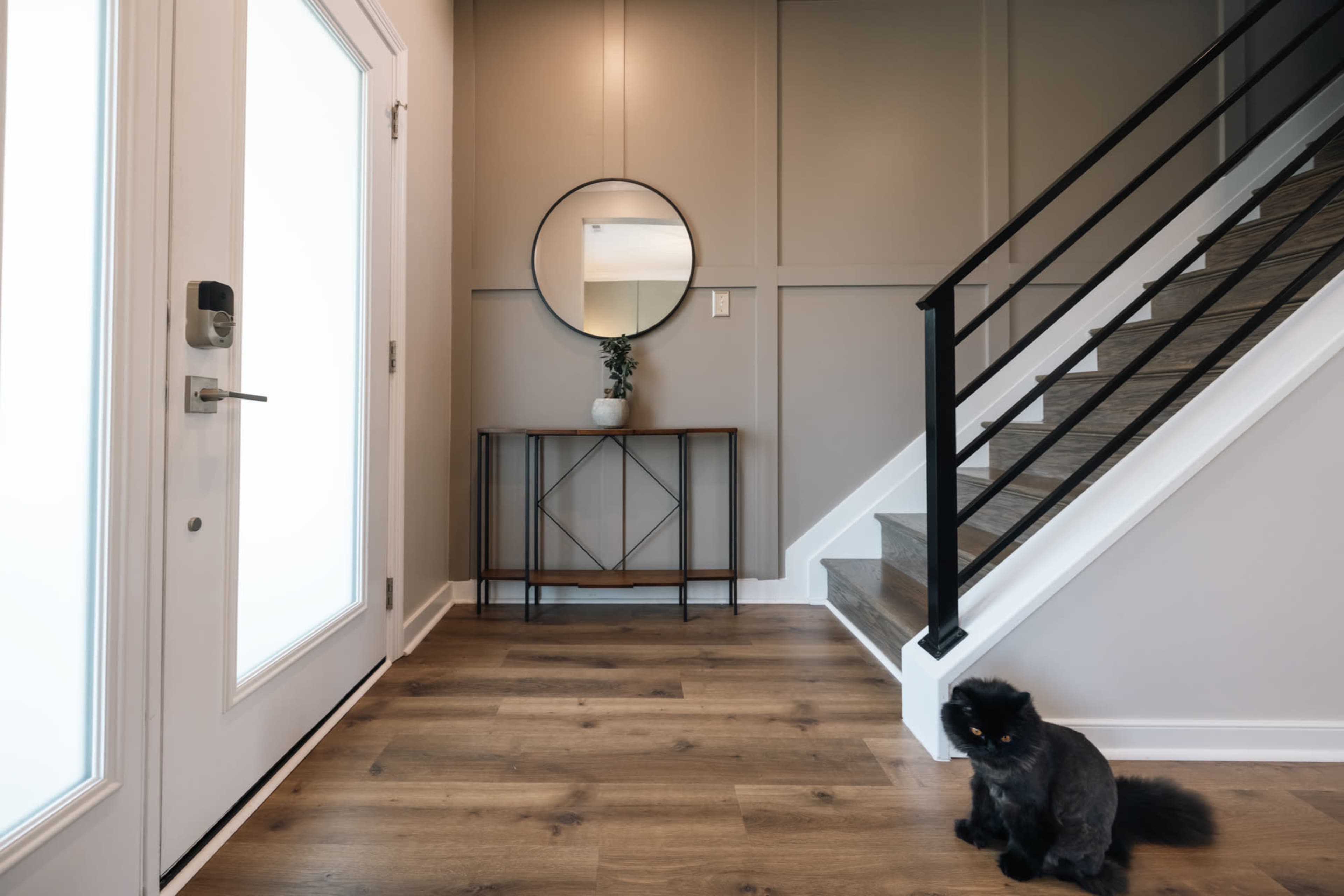 The image shows a modern entryway featuring a white door, a round mirror above a wooden console table, and a black cat sitting on the floor near a staircase.