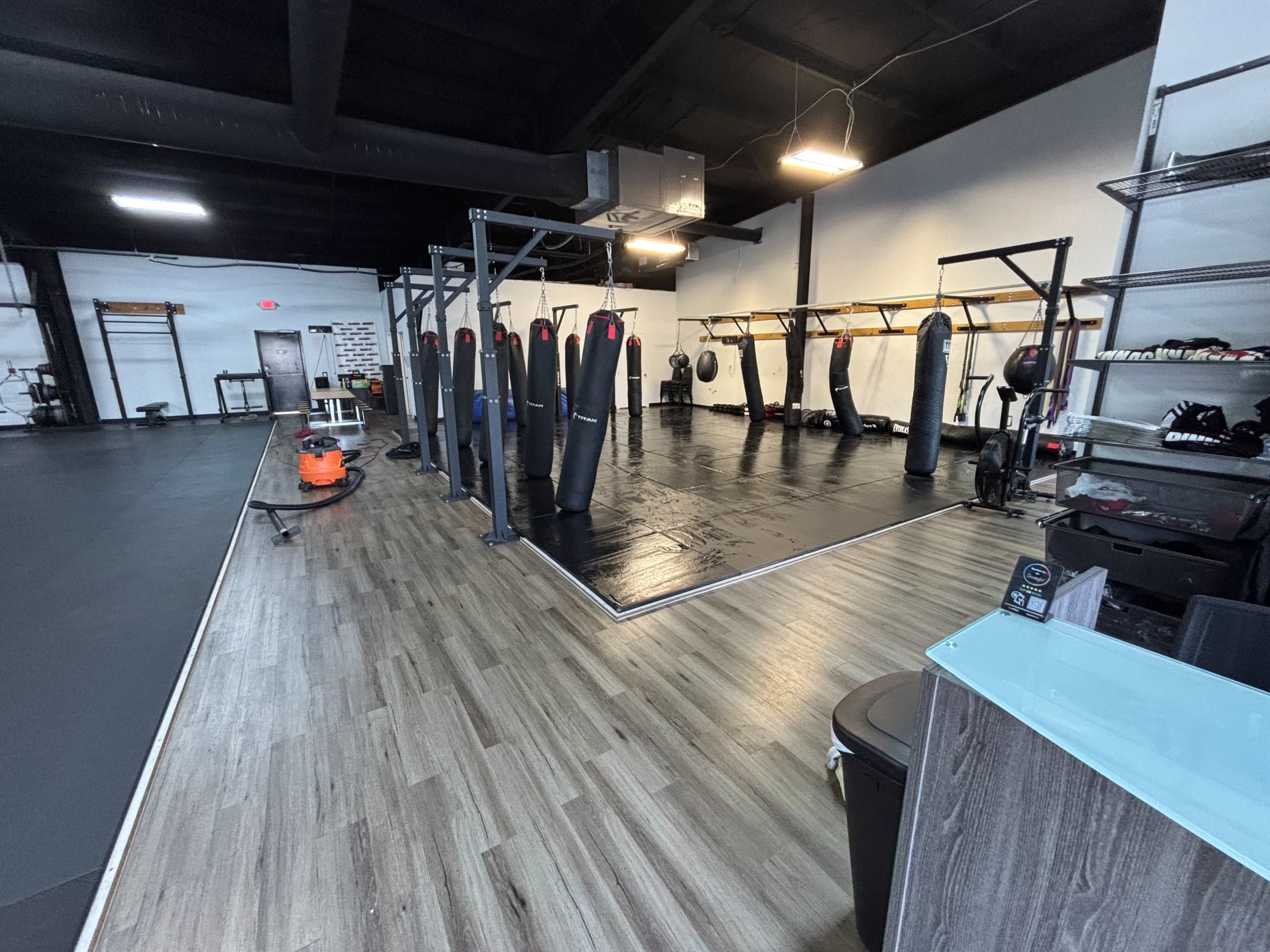 The image shows a spacious gym interior featuring several upright punching bags along a black training area, with wooden flooring and gym equipment visible in the background.