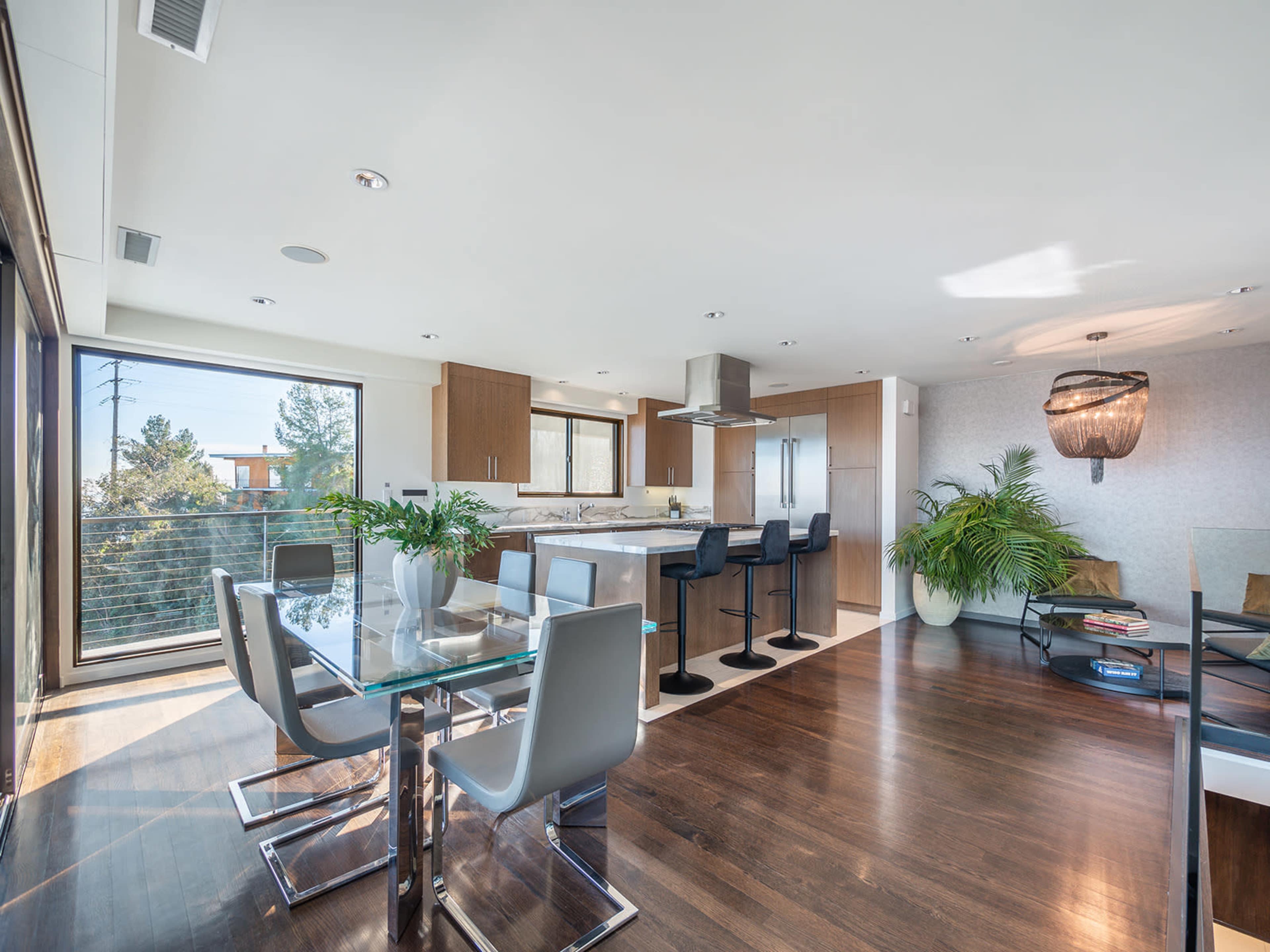 The image shows a modern kitchen and dining area with a glass dining table, sleek chairs, and large windows that provide natural light.