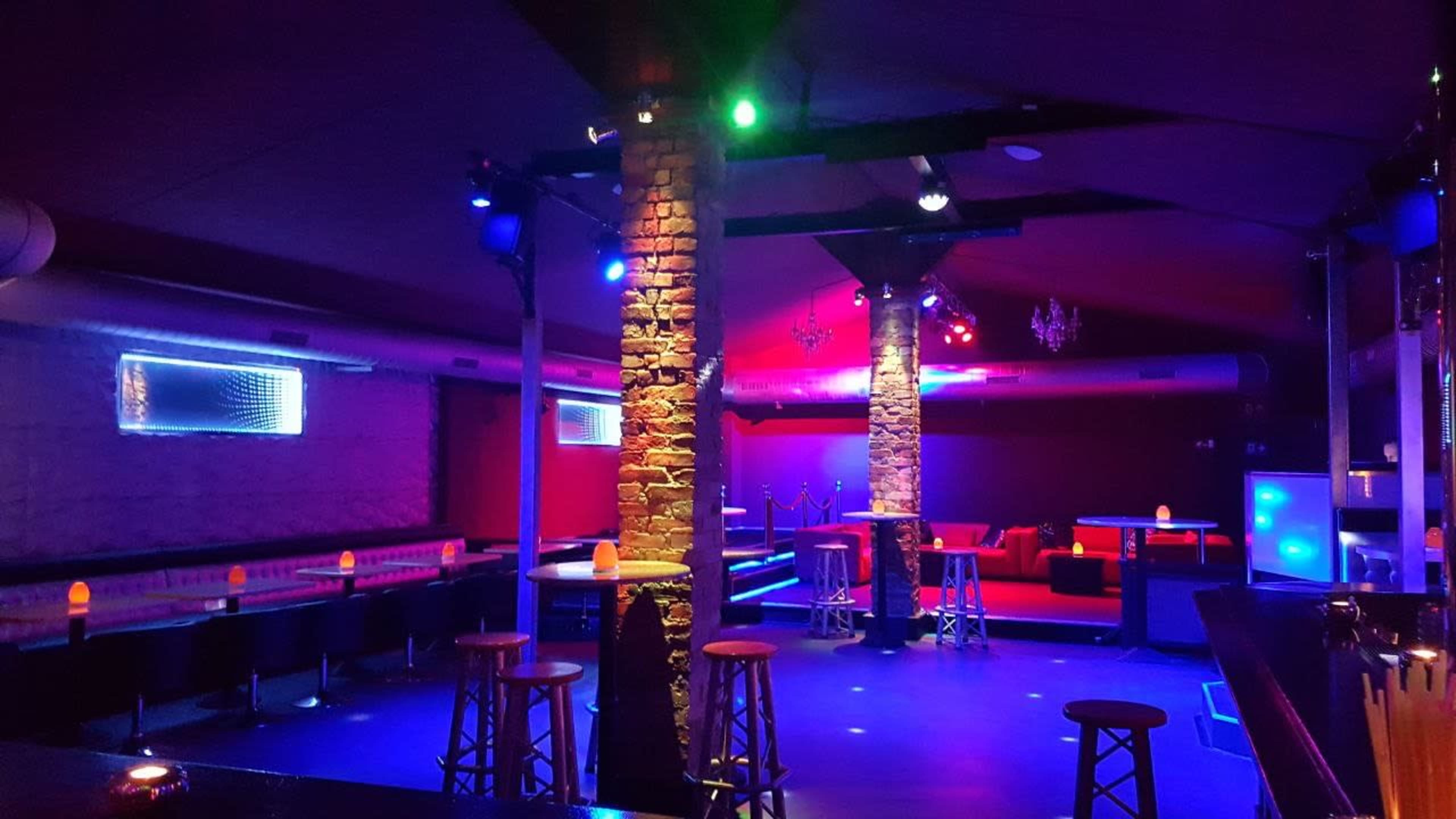 The interior of a dimly lit lounge features stone pillars, bar stools, and colorful lights illuminating the space.