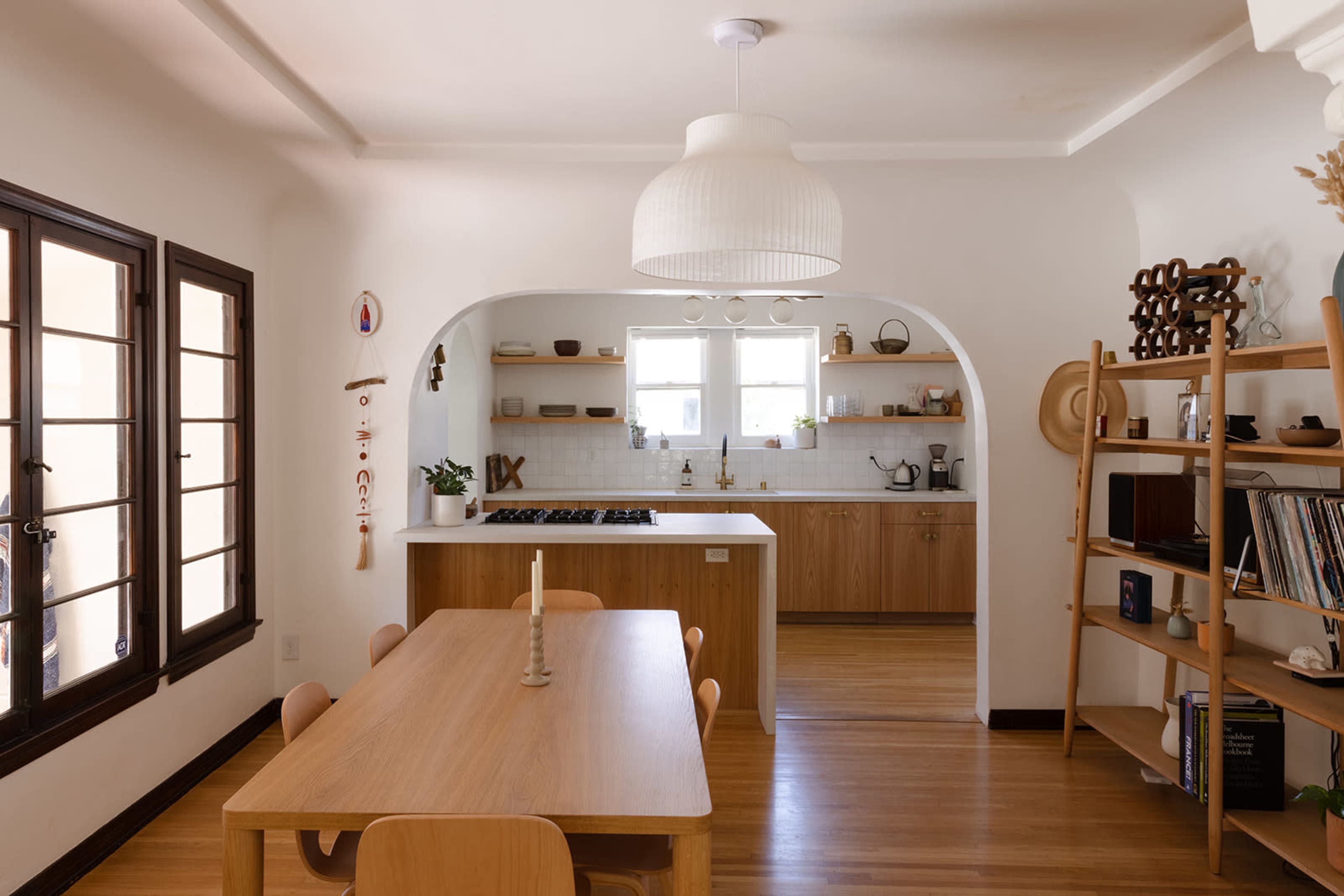 A modern dining area connected to a kitchen, featuring a wooden table with chairs, an archway leading to the kitchen, and shelves displaying kitchenware.