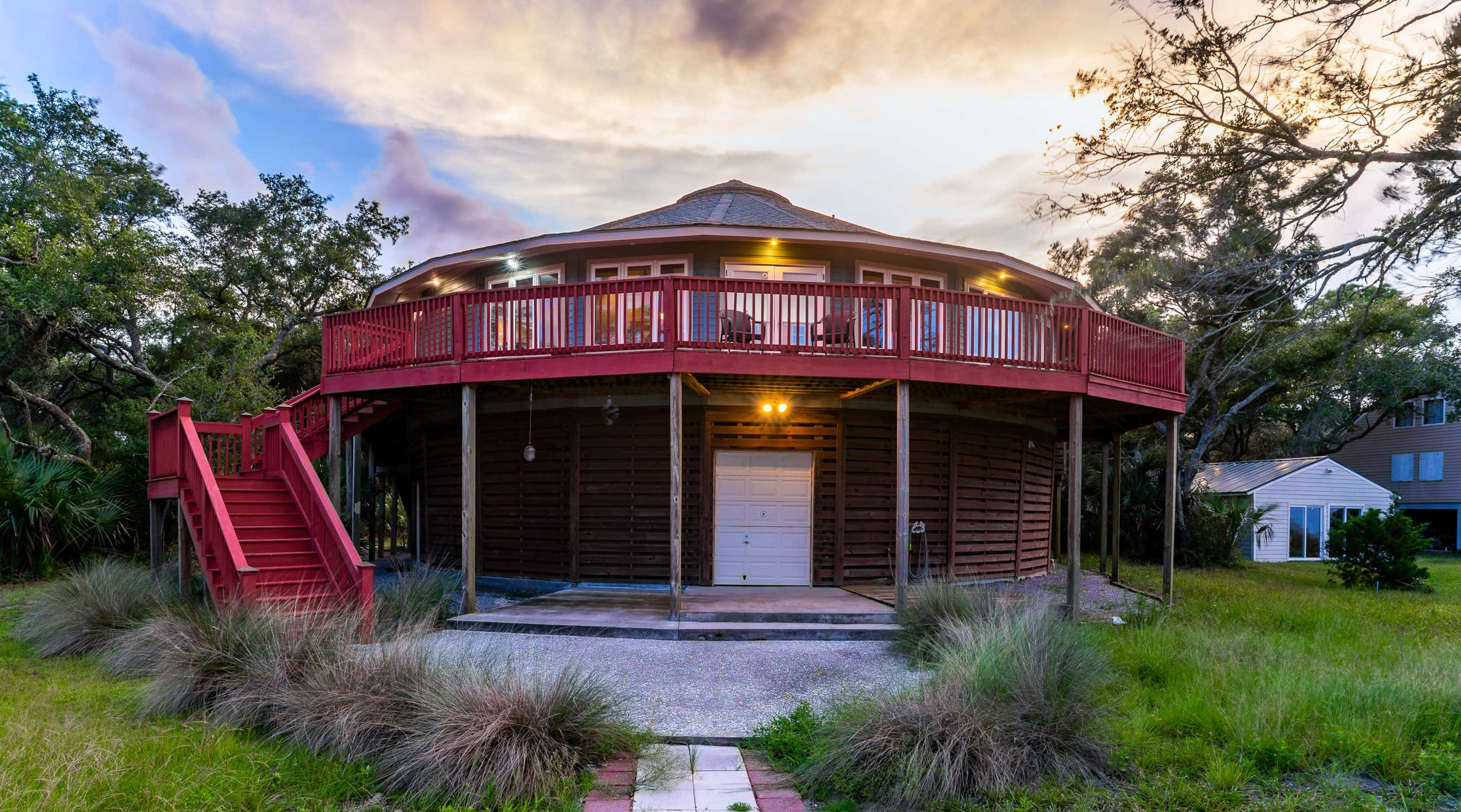 A circular wooden house with a red balcony is surrounded by grass and trees under a cloudy sky.
