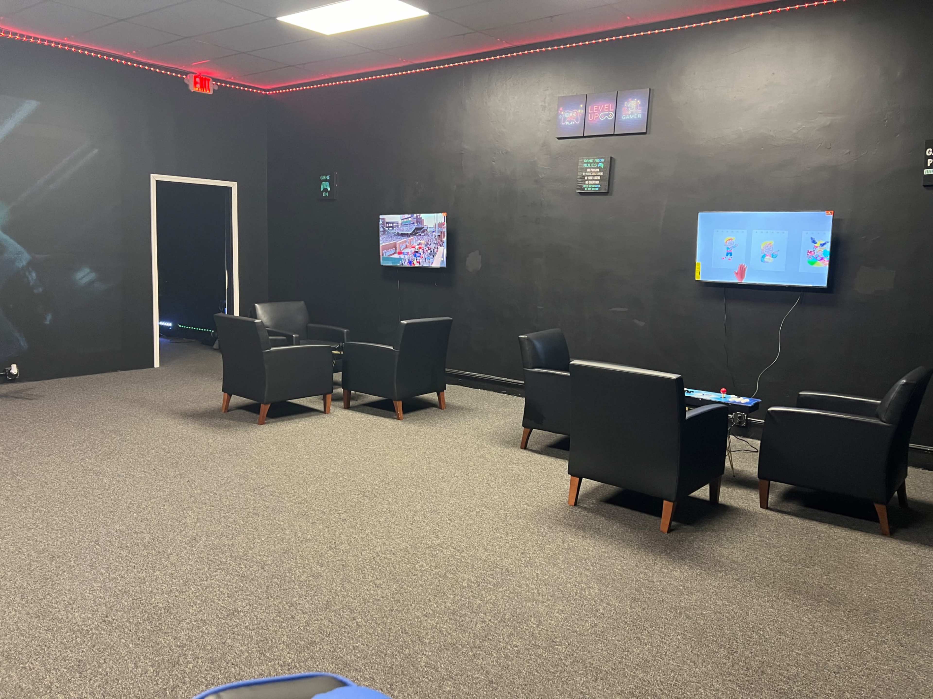 A room with dark walls, two televisions mounted on the wall, and four black chairs arranged in a seating area with carpeted flooring.