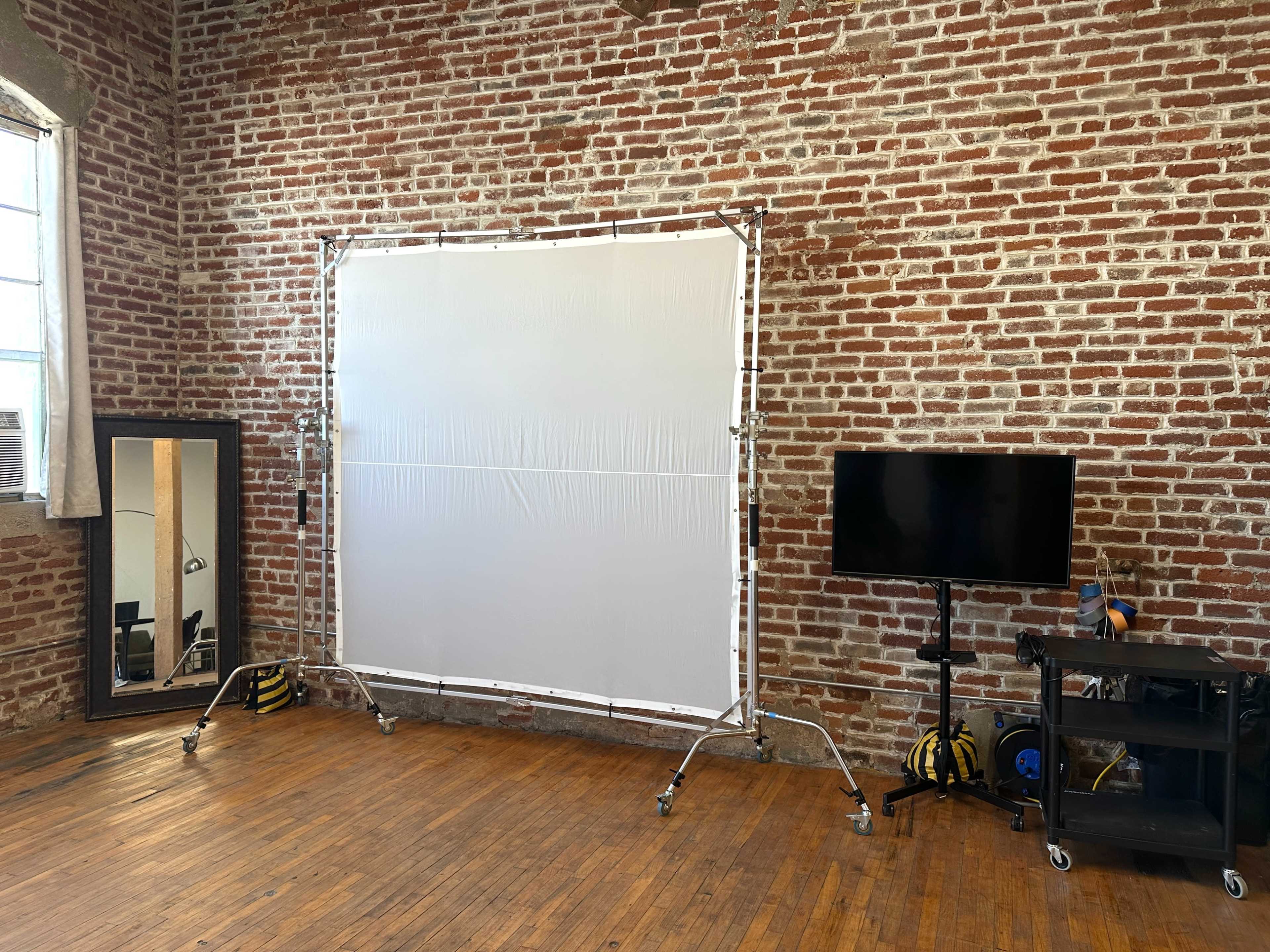 The image shows a studio space with a brick wall, featuring a large white backdrop on a frame, a full-length mirror, and a television on a black cart.