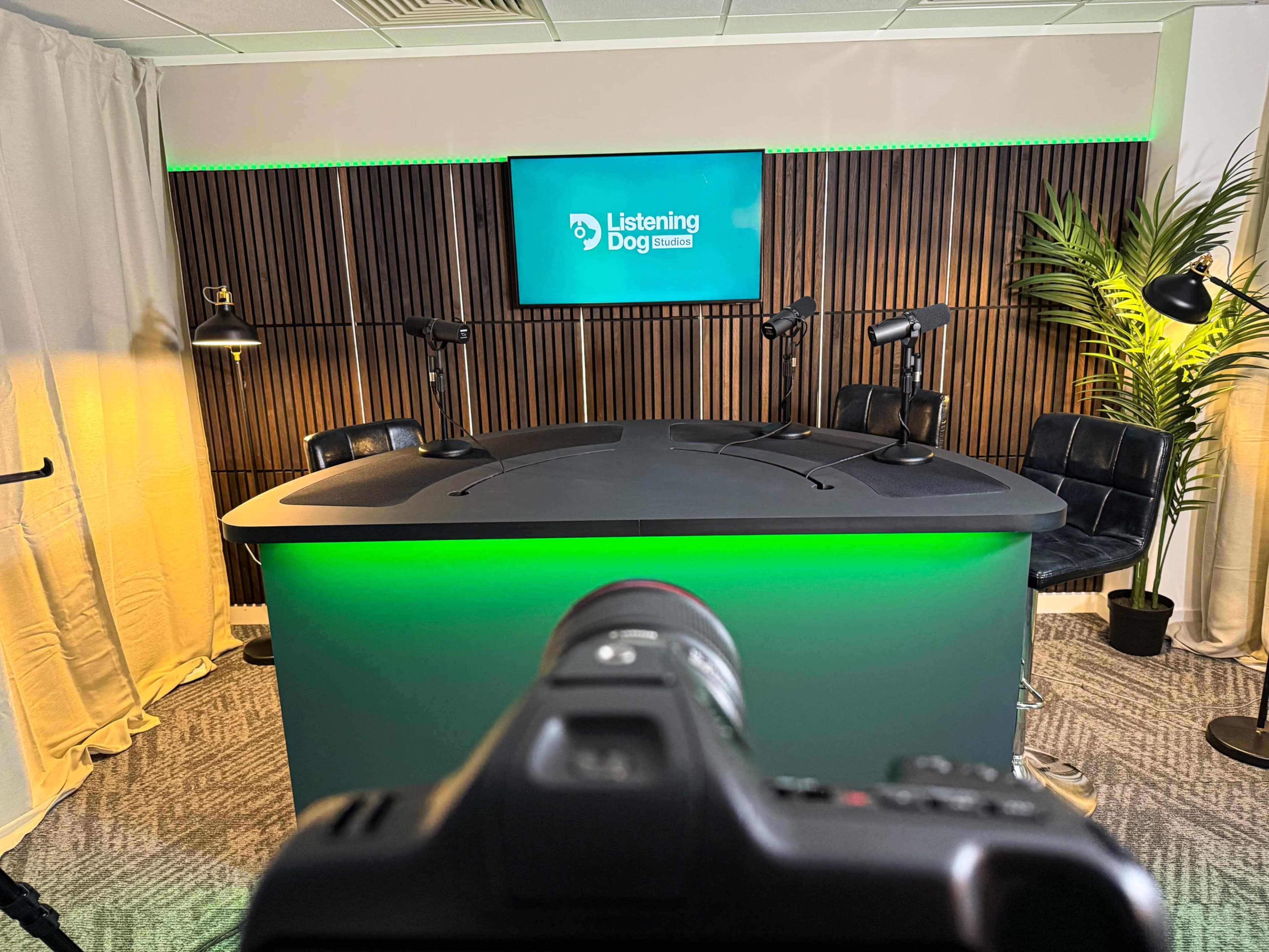 The image shows a modern podcast studio featuring a large table with multiple microphones, a green backlit panel displaying the name "Listening Dog," and a few plants in the background.
