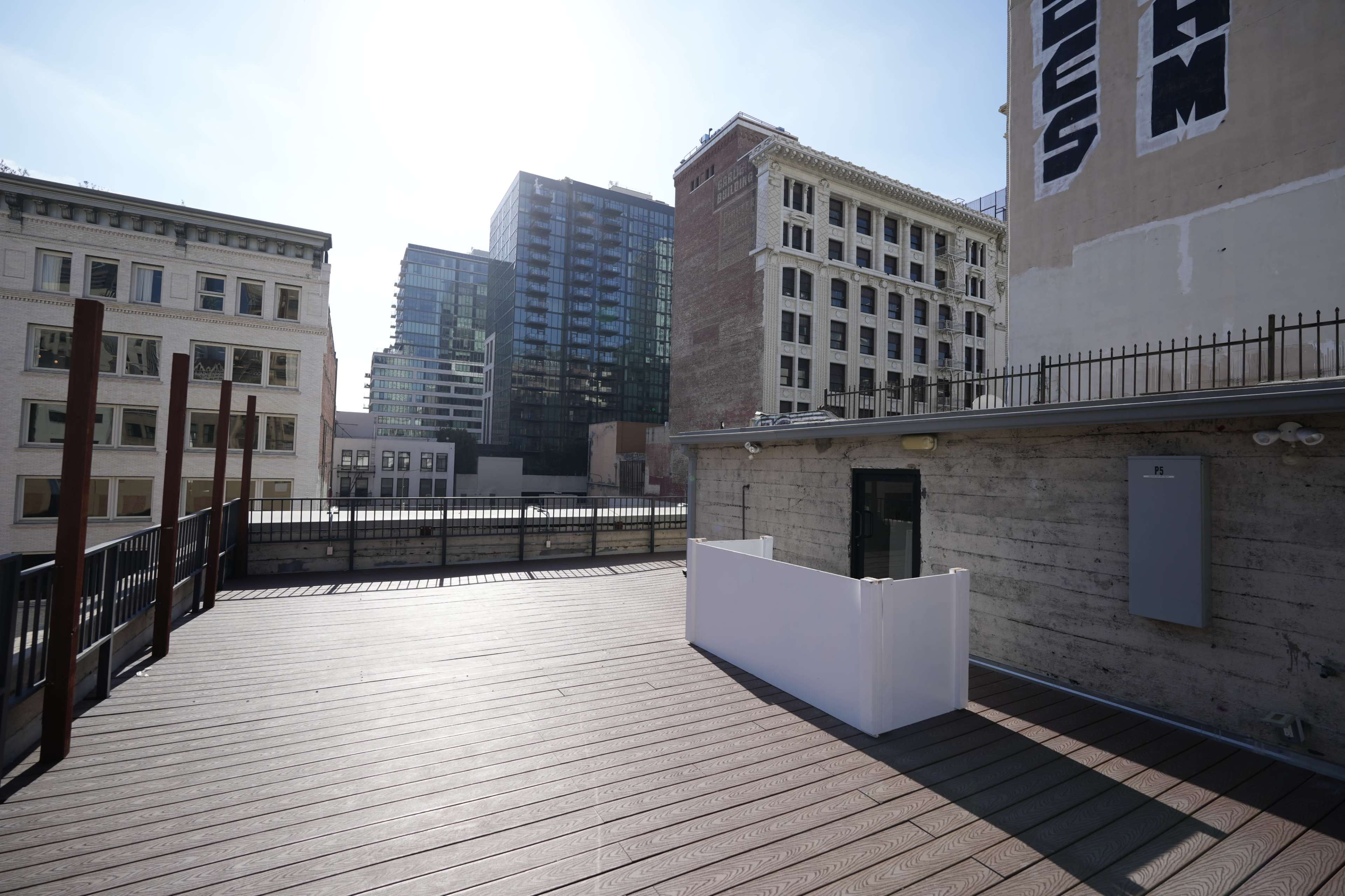 The image shows a rooftop terrace with wooden flooring, surrounded by tall buildings in an urban setting.