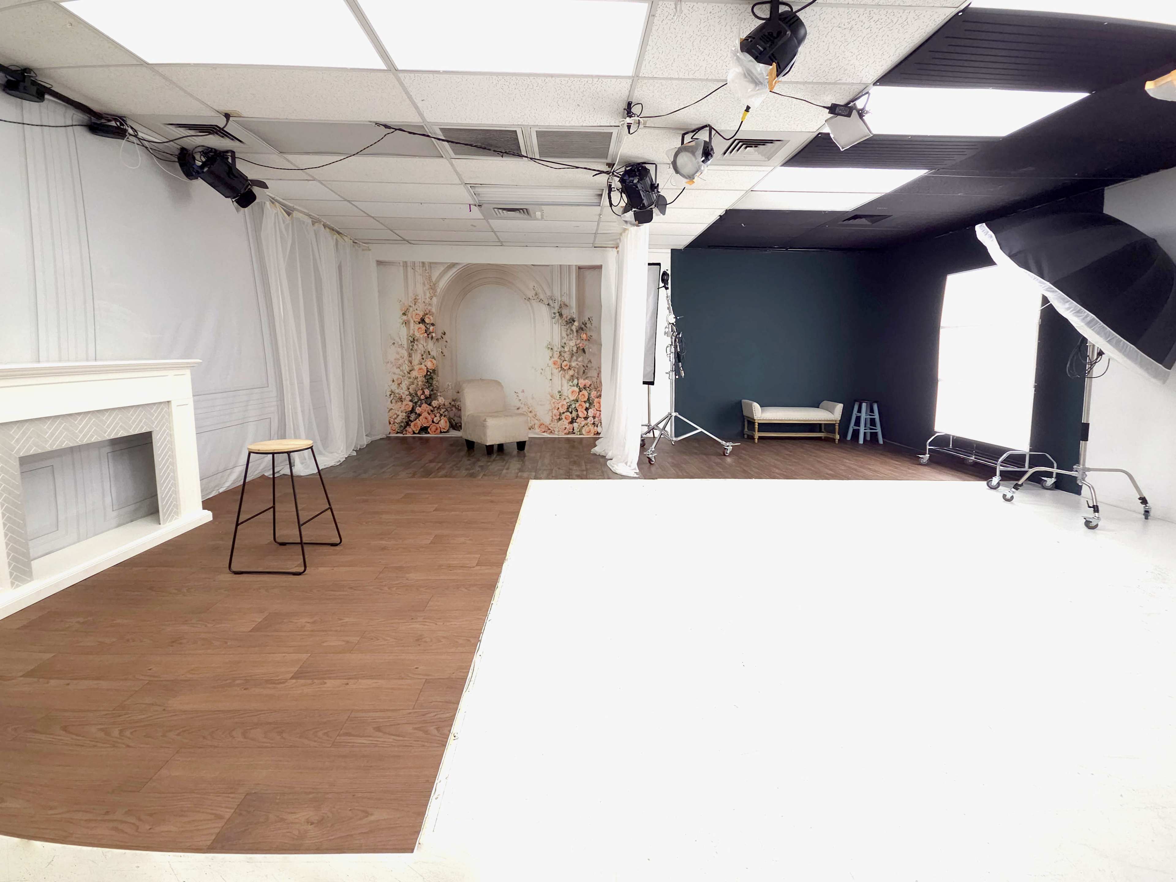 The image depicts a photography studio with a split design featuring a white section, a wooden floor, a decorative backdrop with floral designs, and various lighting equipment.
