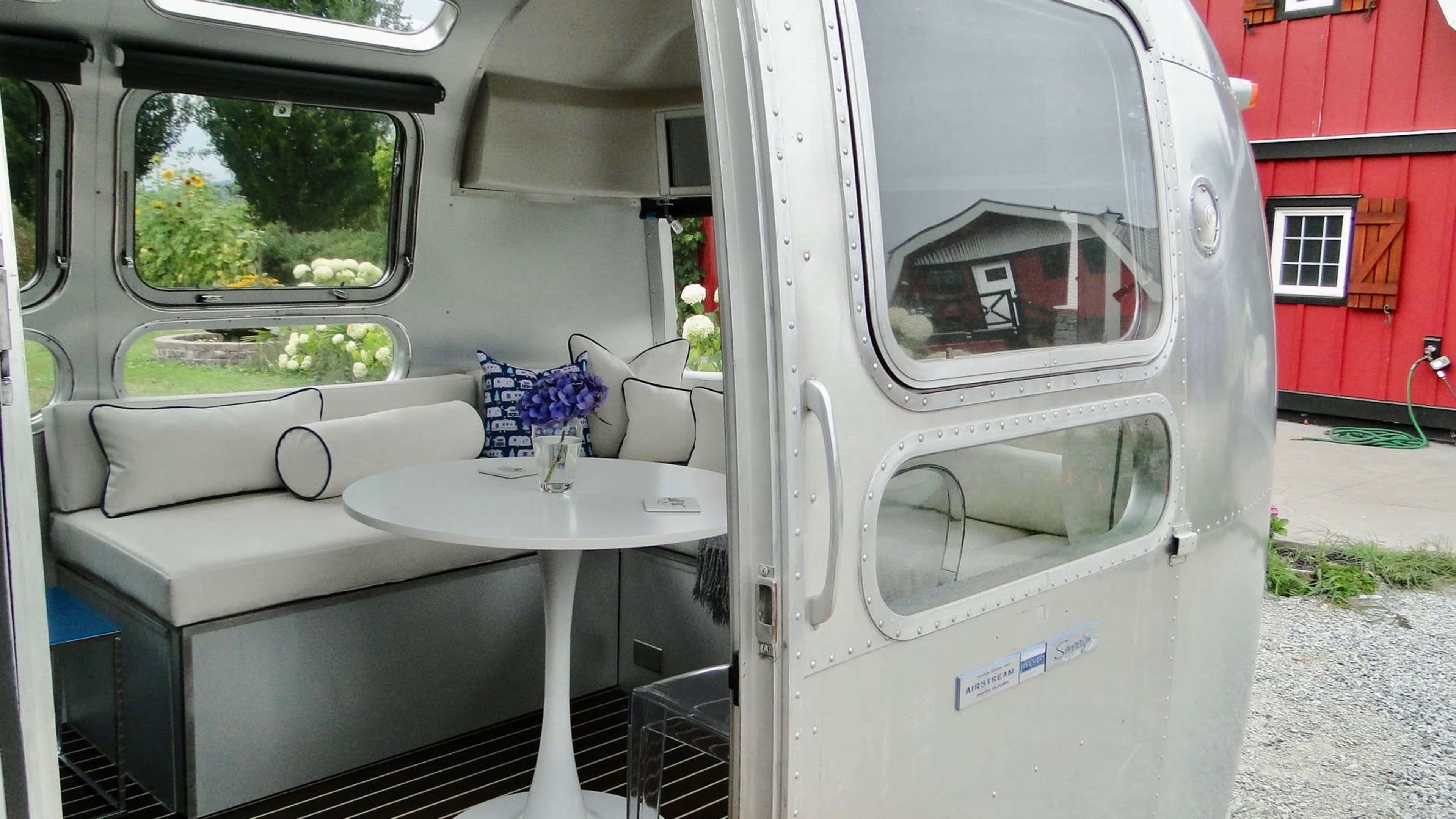 The image shows the interior of an Airstream trailer with a circular table and a seating area that includes cushions, along with a view of a red building outside.