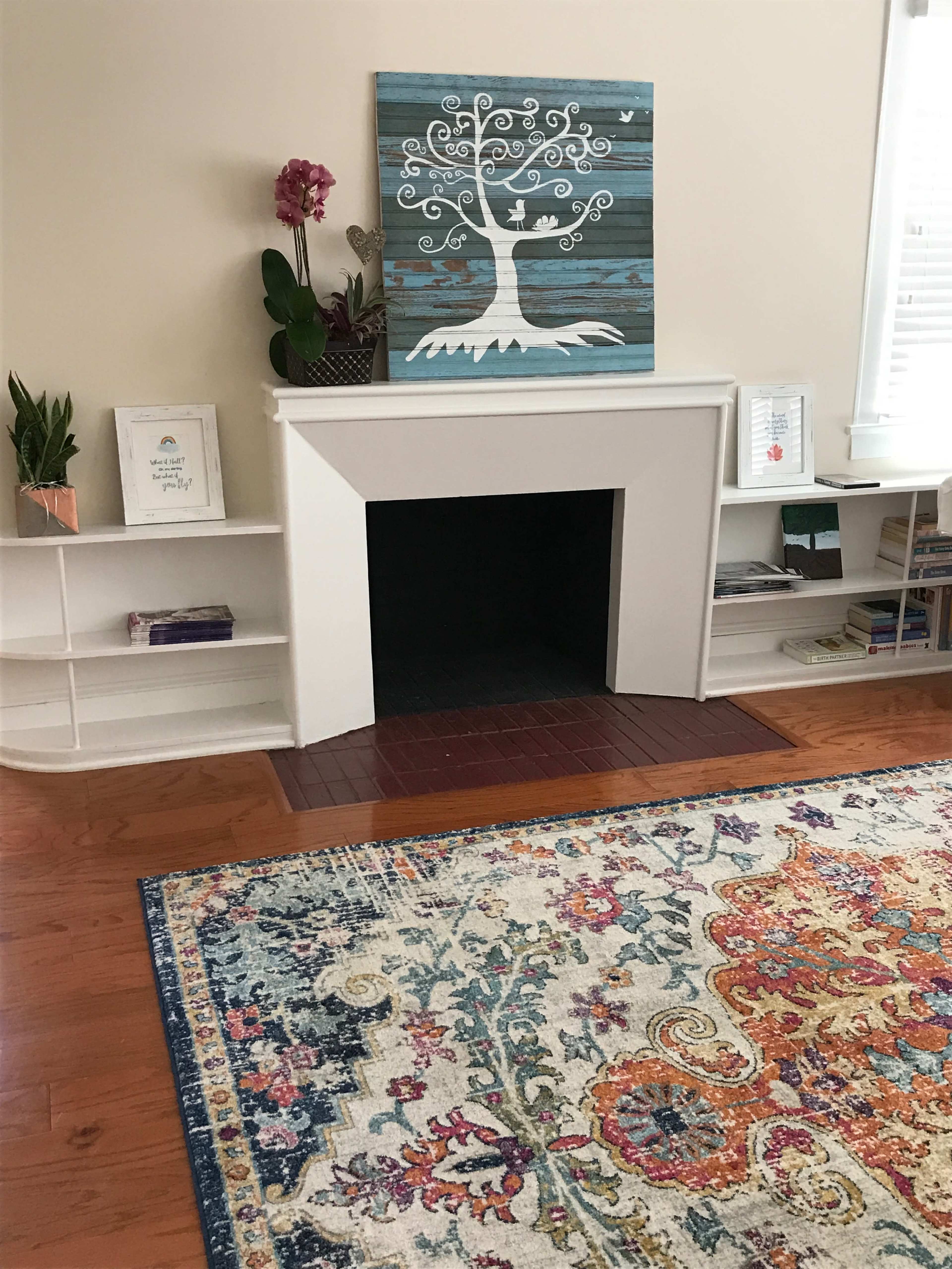 A living room featuring a white fireplace surrounded by built-in shelves, a colorful area rug, and a wall-mounted artwork of a tree.
