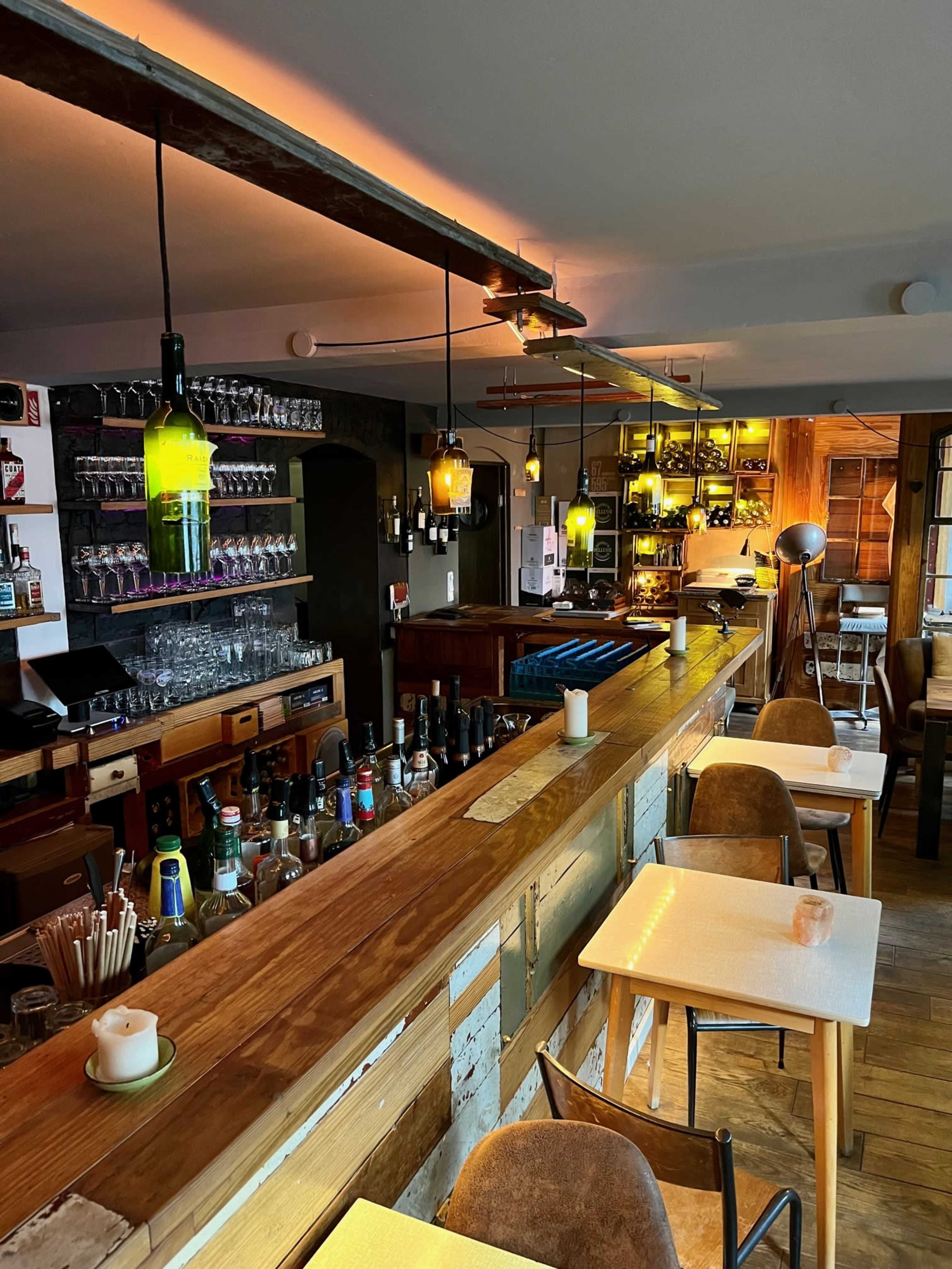 The interior of a bar featuring a wooden counter, various bottles on shelves, and pendant lights illuminating the space.