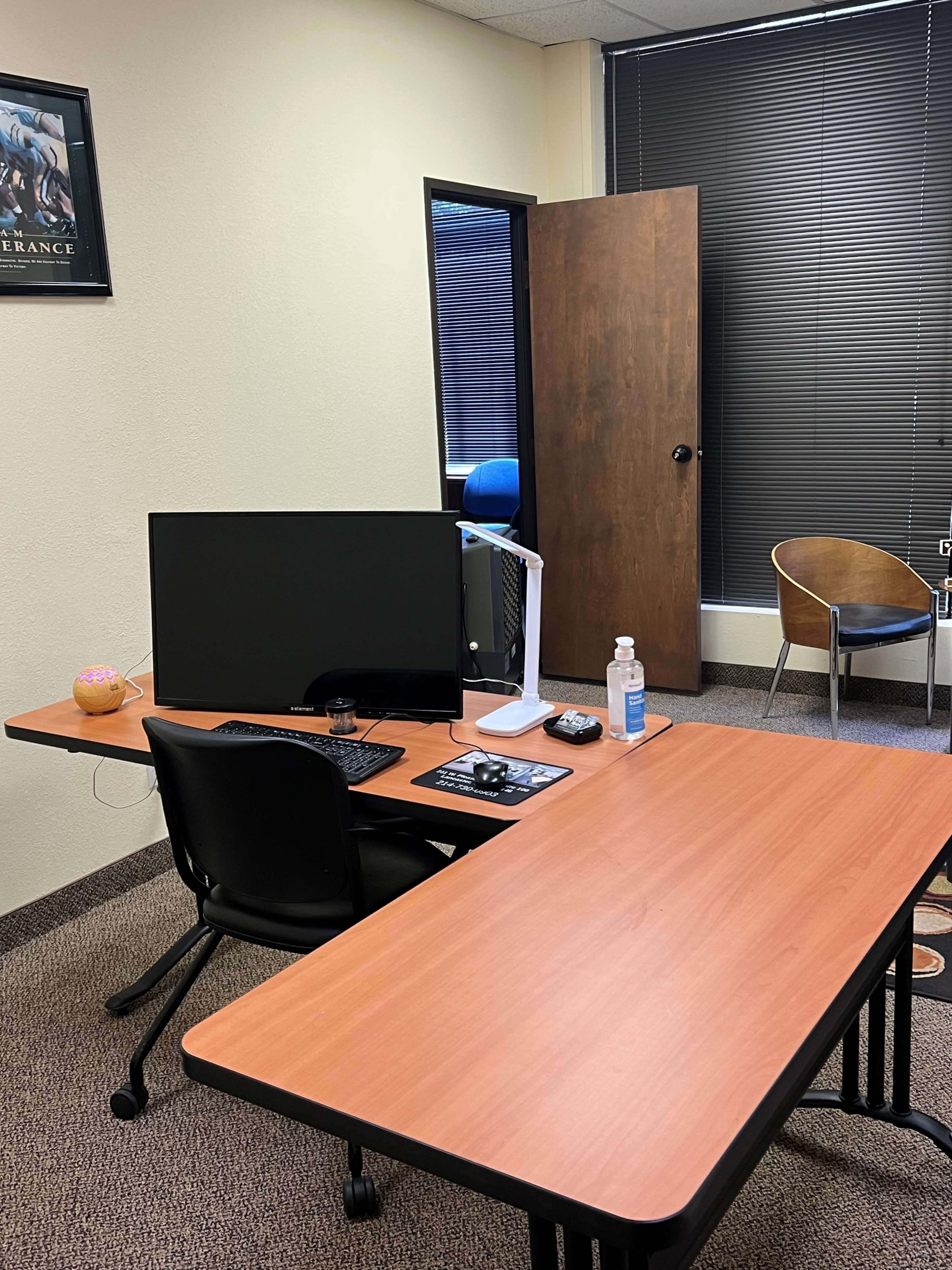 The image shows a small office with a desk featuring a computer monitor, keyboard, and a mouse, alongside a chair and an open door leading to a hallway.