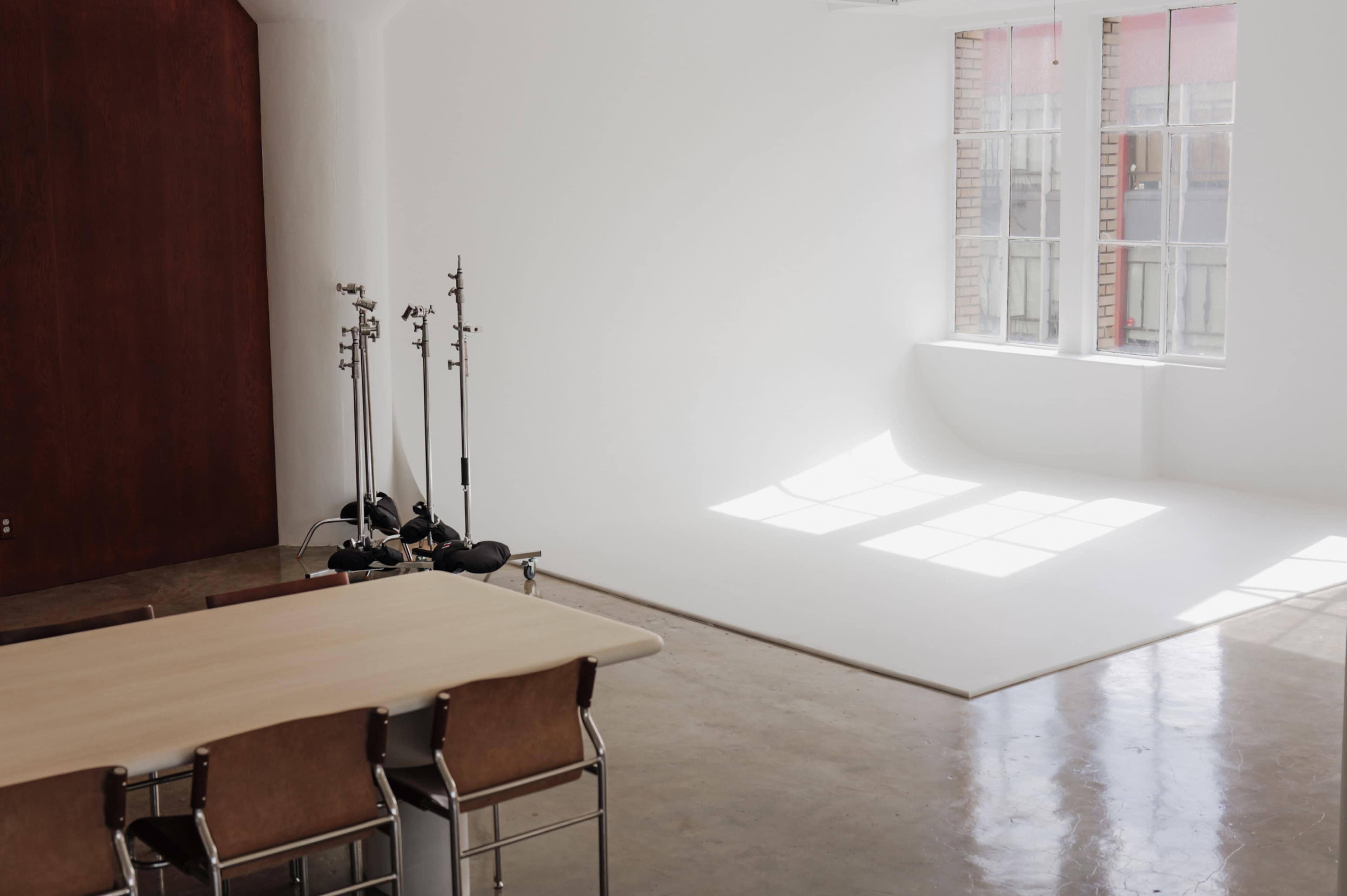 The image shows a spacious, well-lit studio with a wooden table and chairs, equipment stands, and large windows allowing natural light to illuminate a white backdrop.