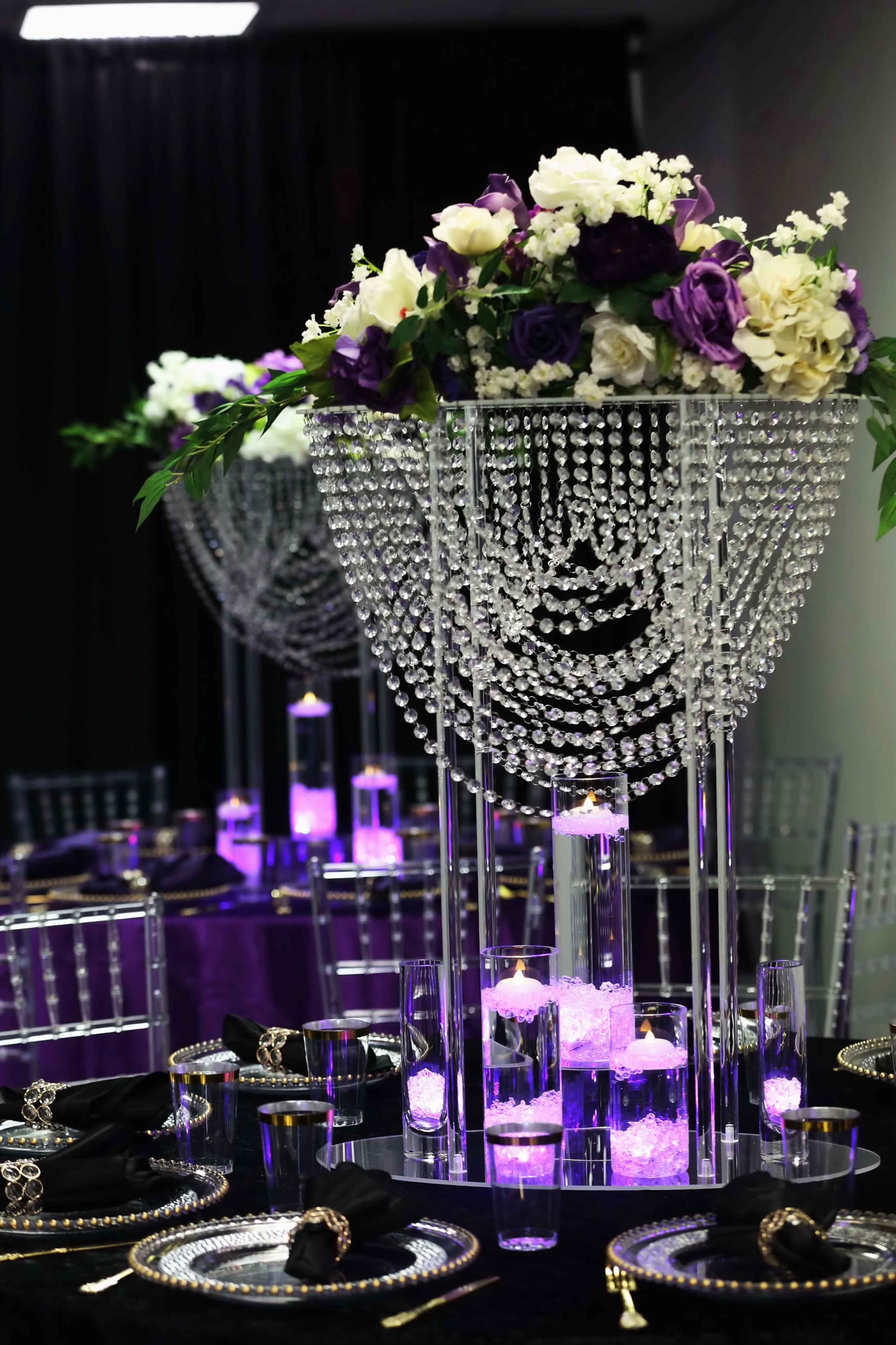 A lavish event setup features tall, crystal-adorned centerpieces filled with flowers and illuminated candles on elegantly set black tables.