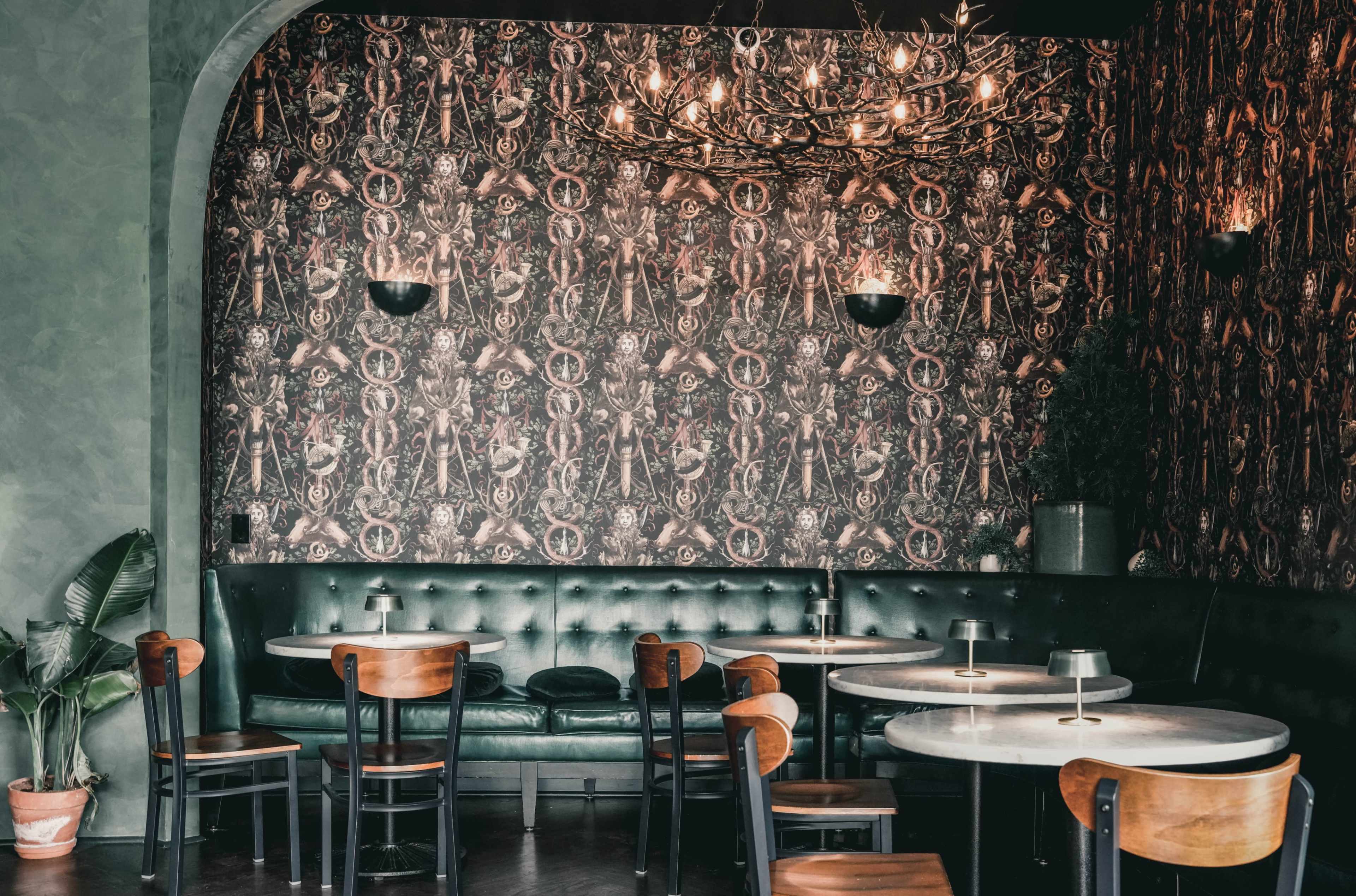 The image shows a restaurant interior featuring a green wall, patterned wallpaper, dark seating, and round marble tables.