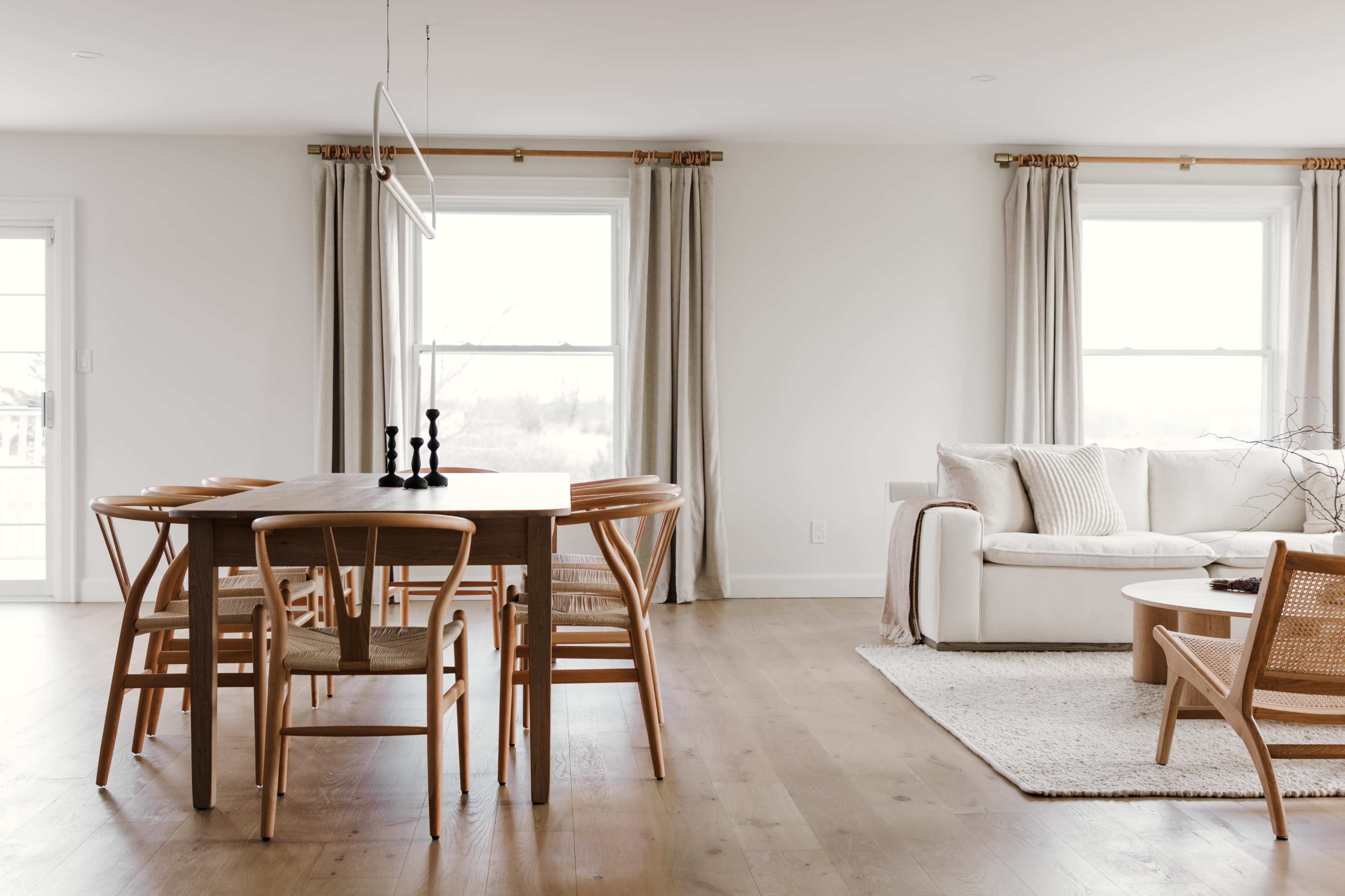 A modern dining area with a wooden table surrounded by six chairs, a sofa, and large windows draped with light curtains.
