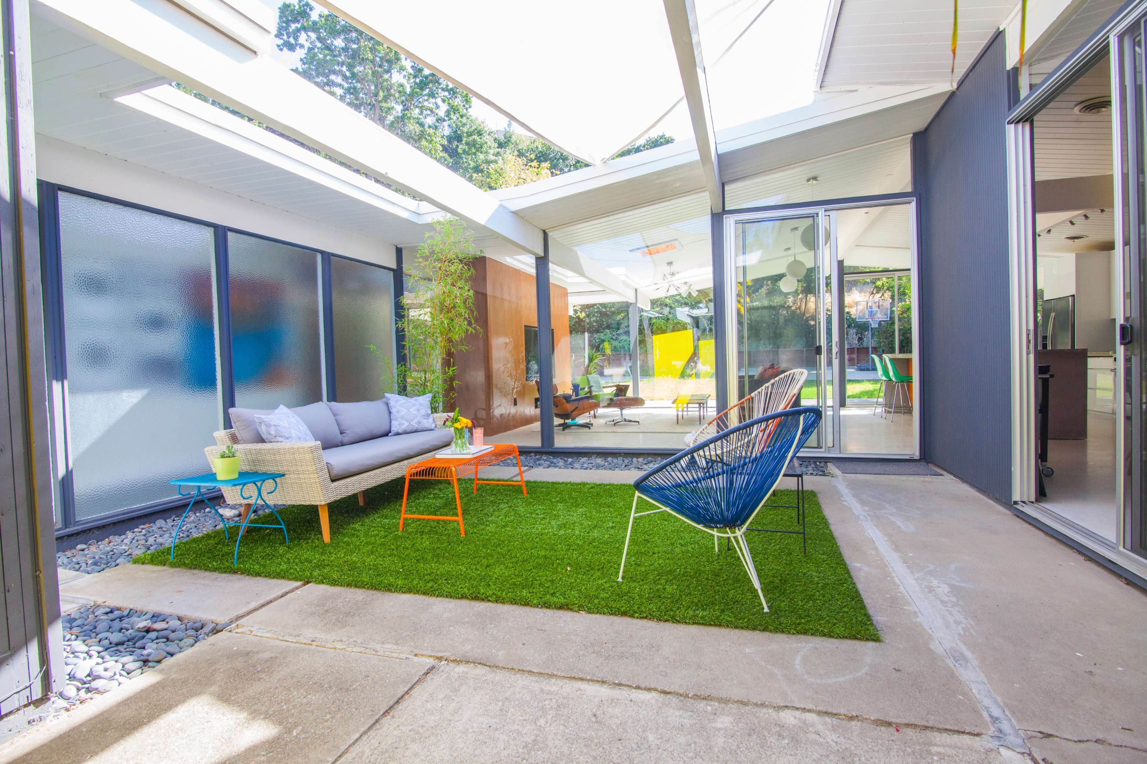 A modern outdoor seating area with artificial grass, featuring a gray sofa, orange side table, and blue chair, surrounded by glass walls and natural light.
