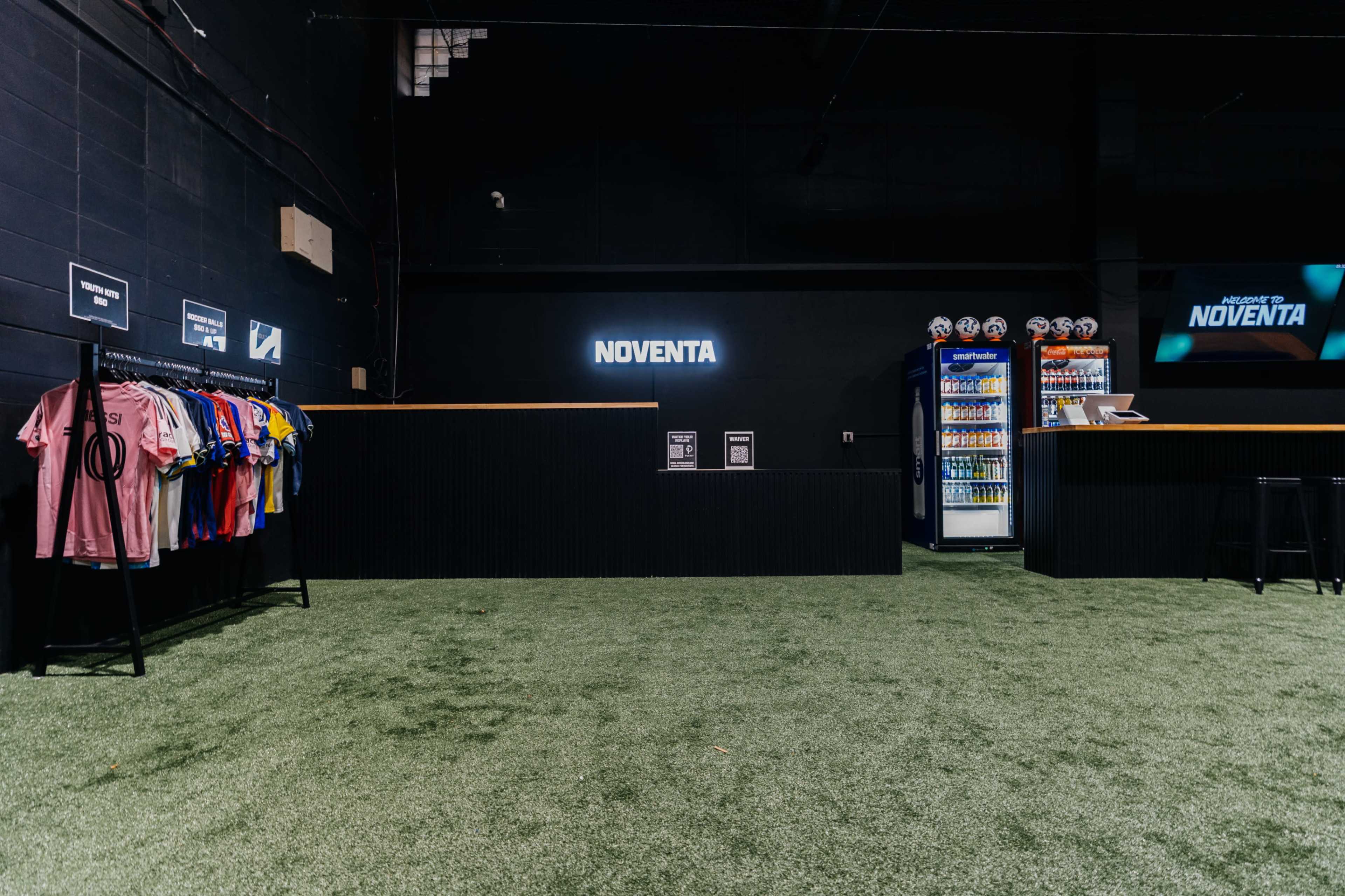 The image shows a retail space with a turf floor, featuring a display of colorful shirts on racks to the left and a vending area with drinks and snacks on the right.