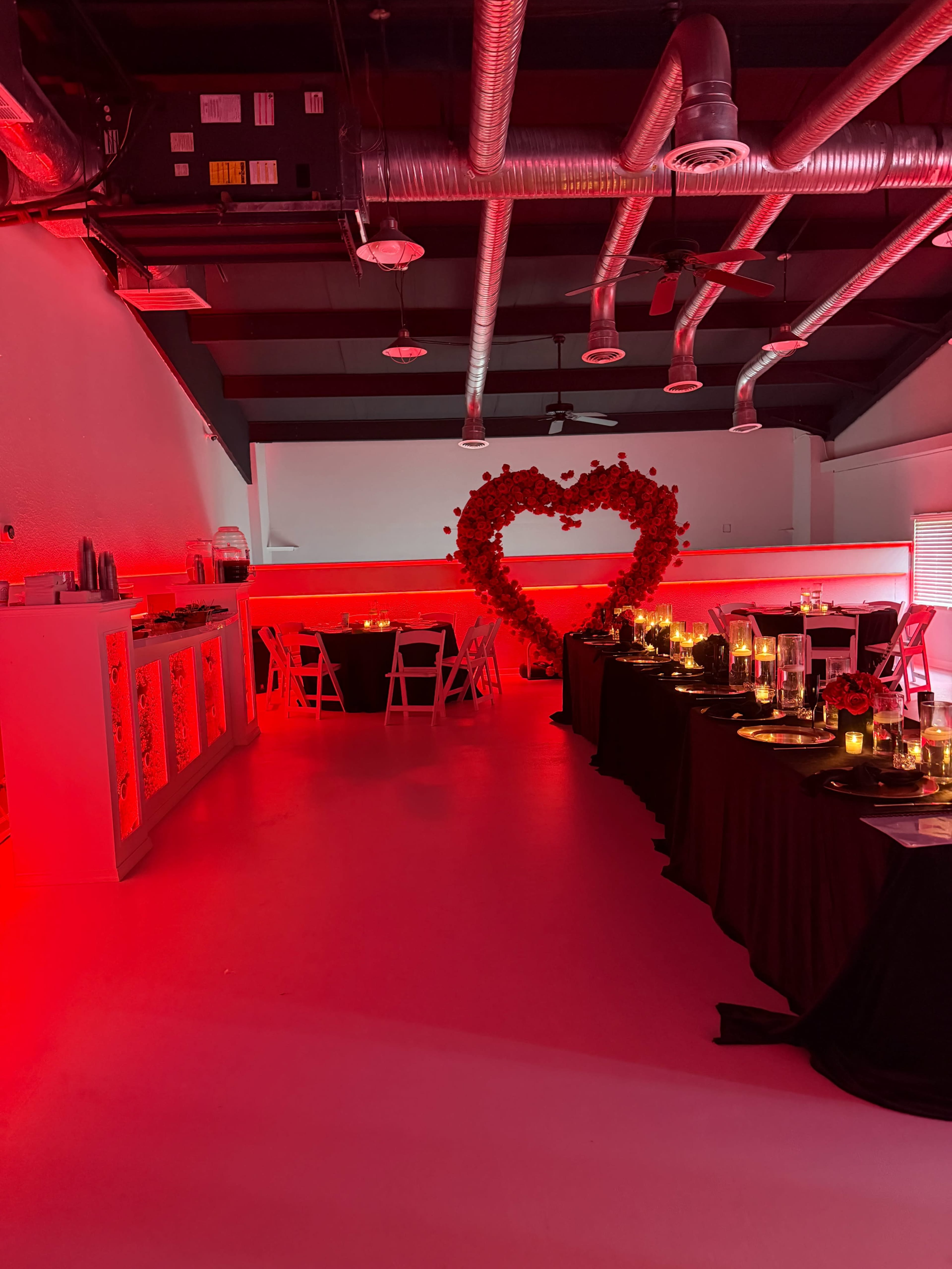 A decorated event space features tables set with candles and red lighting, highlighted by a heart-shaped floral display on the wall.