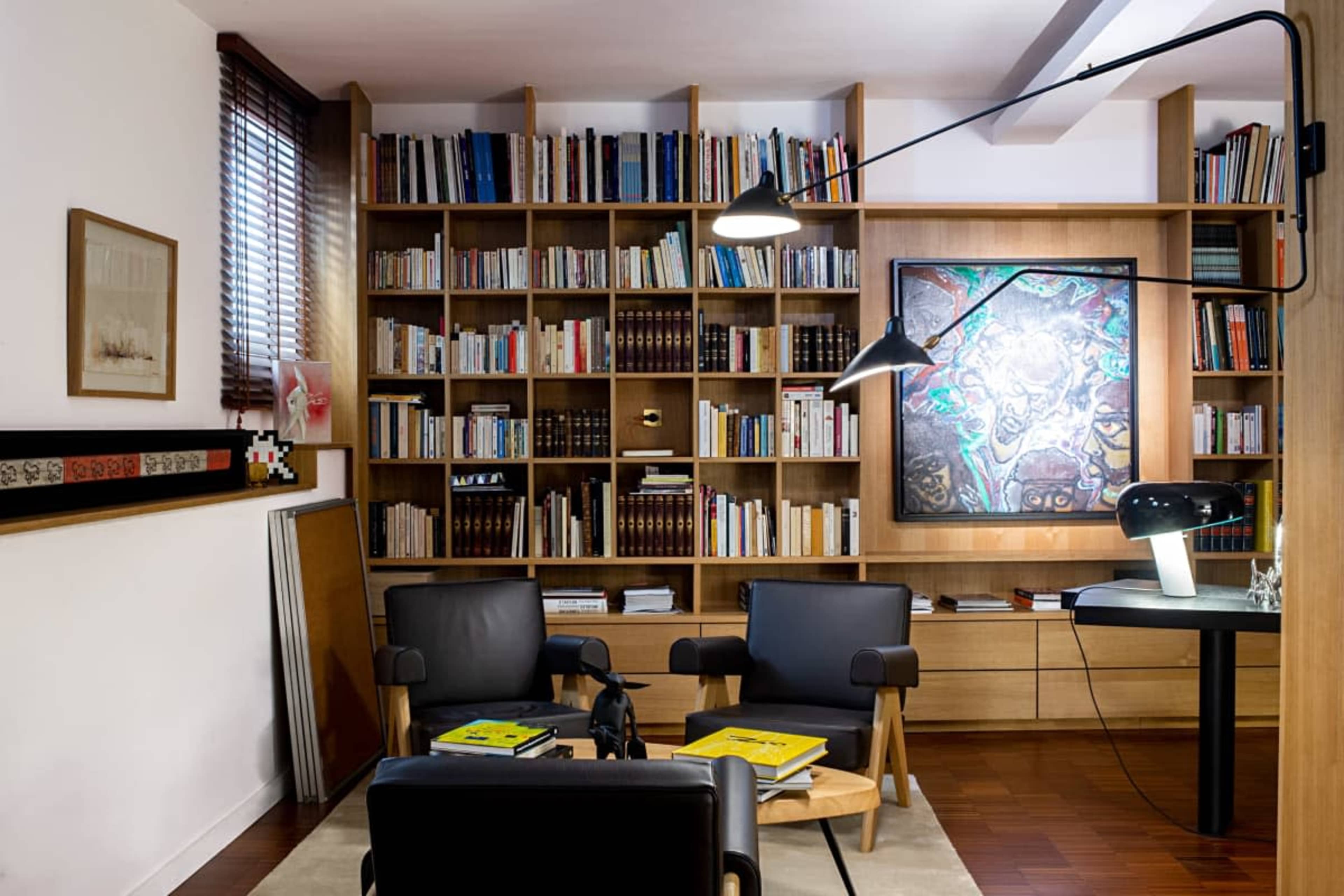 The image shows a modern living space featuring a bookshelf filled with books, two black armchairs, and a table lamp beside a small table.