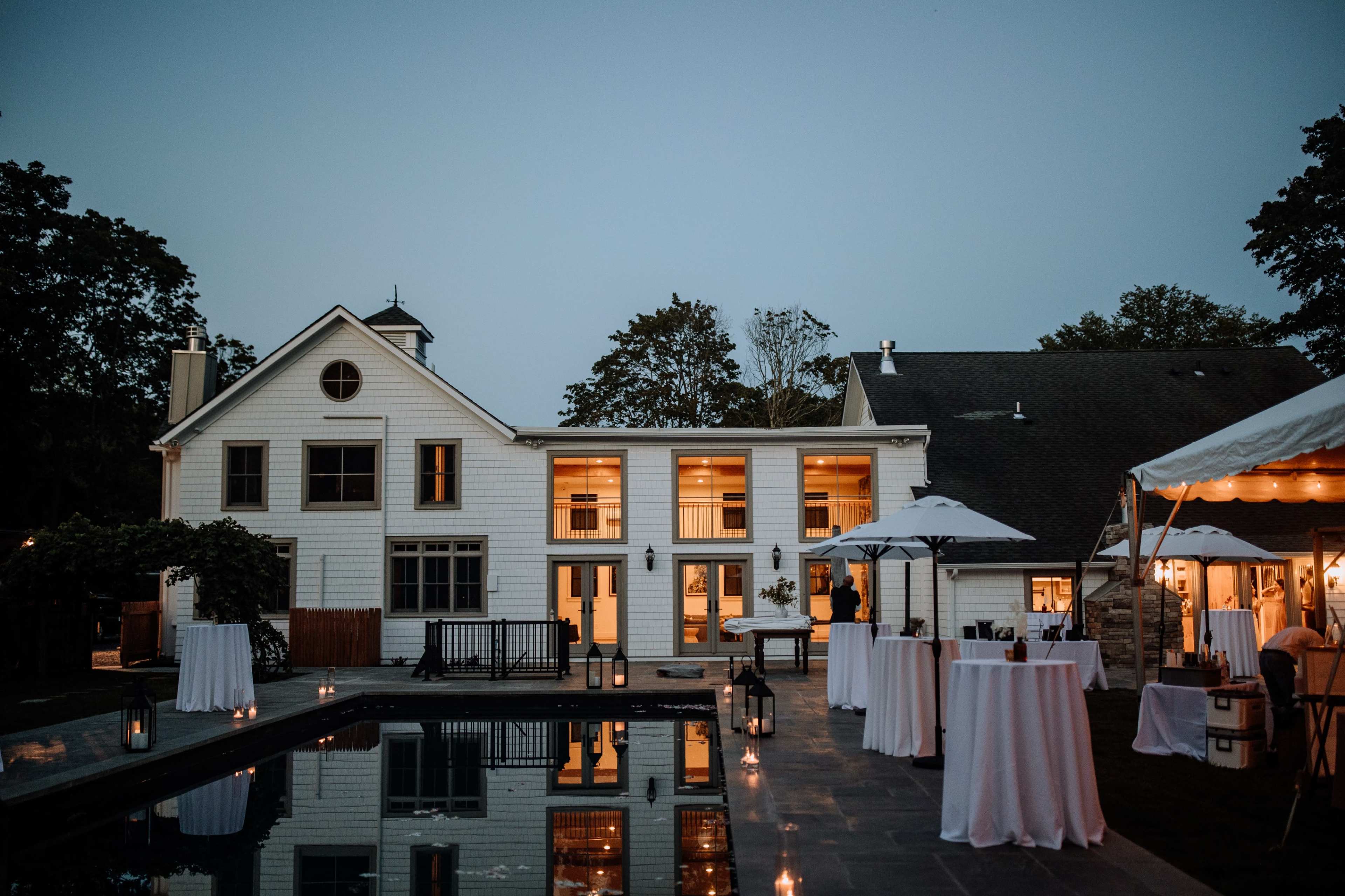 A modern house with large windows and a pool, surrounded by tables under umbrellas set up for an outdoor event during twilight.