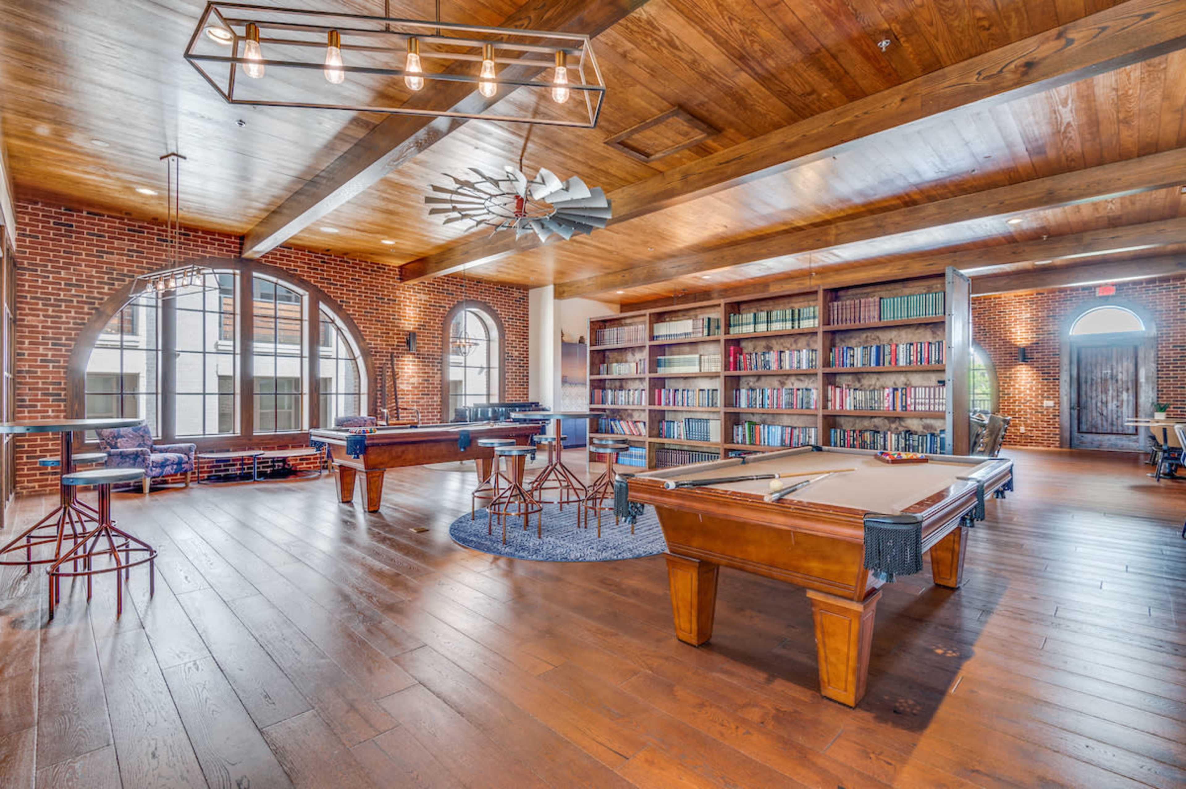 The image depicts a spacious room with wooden floors, exposed brick walls, a pool table, and a bookshelf filled with books, featuring large arched windows that allow natural light to enter.
