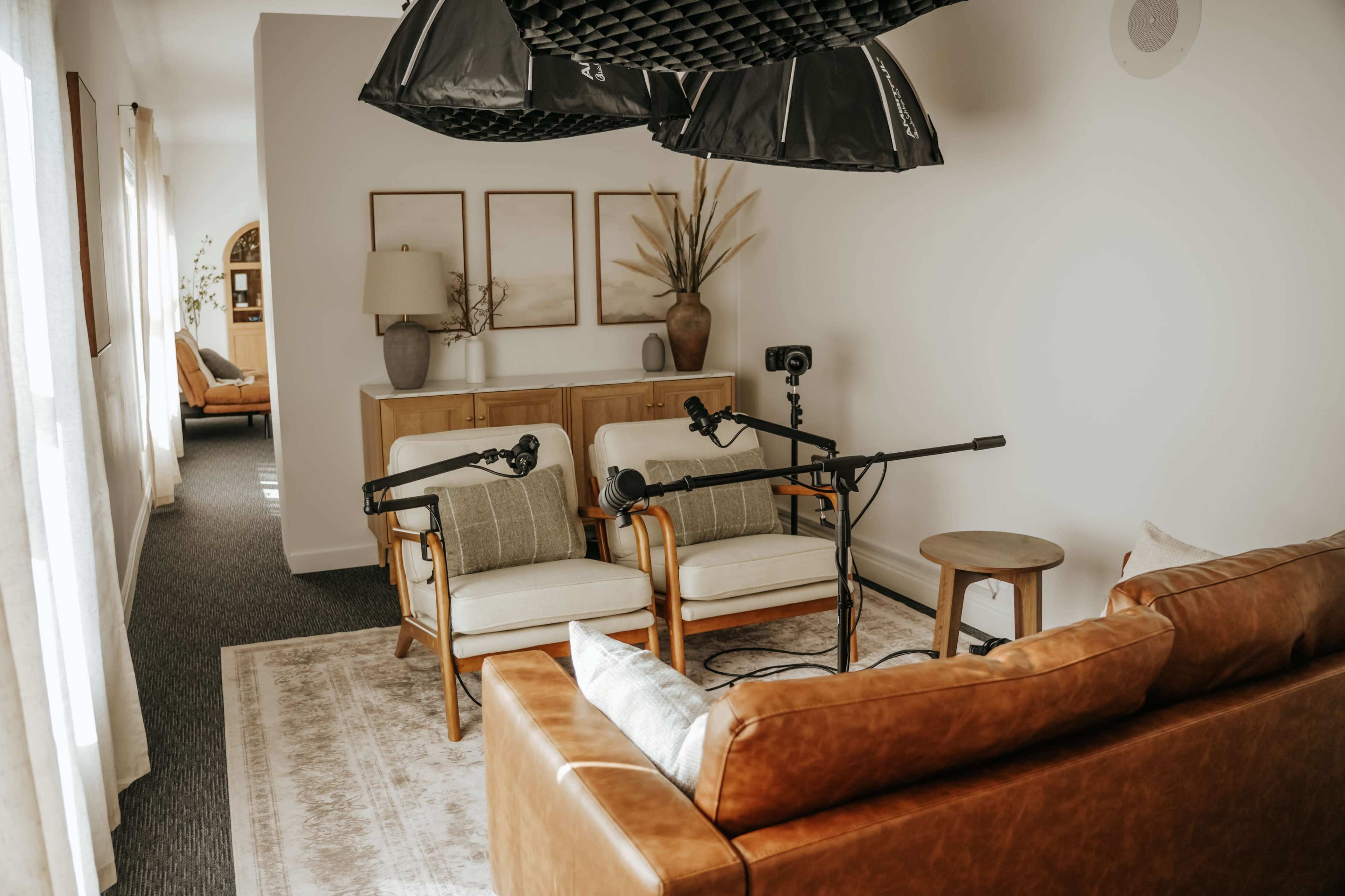 The image shows a cozy living room setup with two armchairs and a leather sofa, along with a coffee table and microphones arranged for a recording session.
