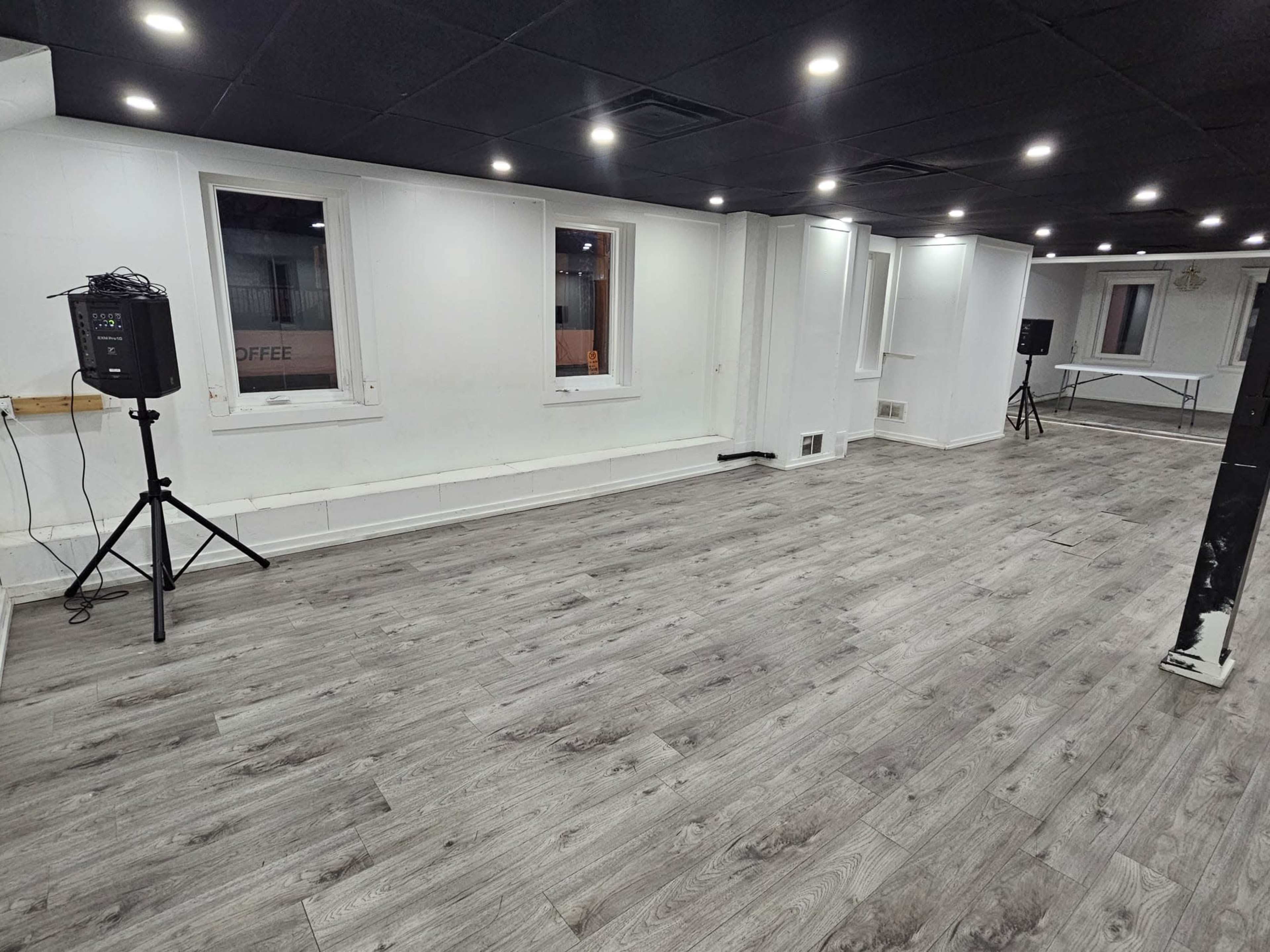 The image shows a spacious, empty room with light-colored wooden flooring, white walls, and several windows, well-lit by overhead lights.