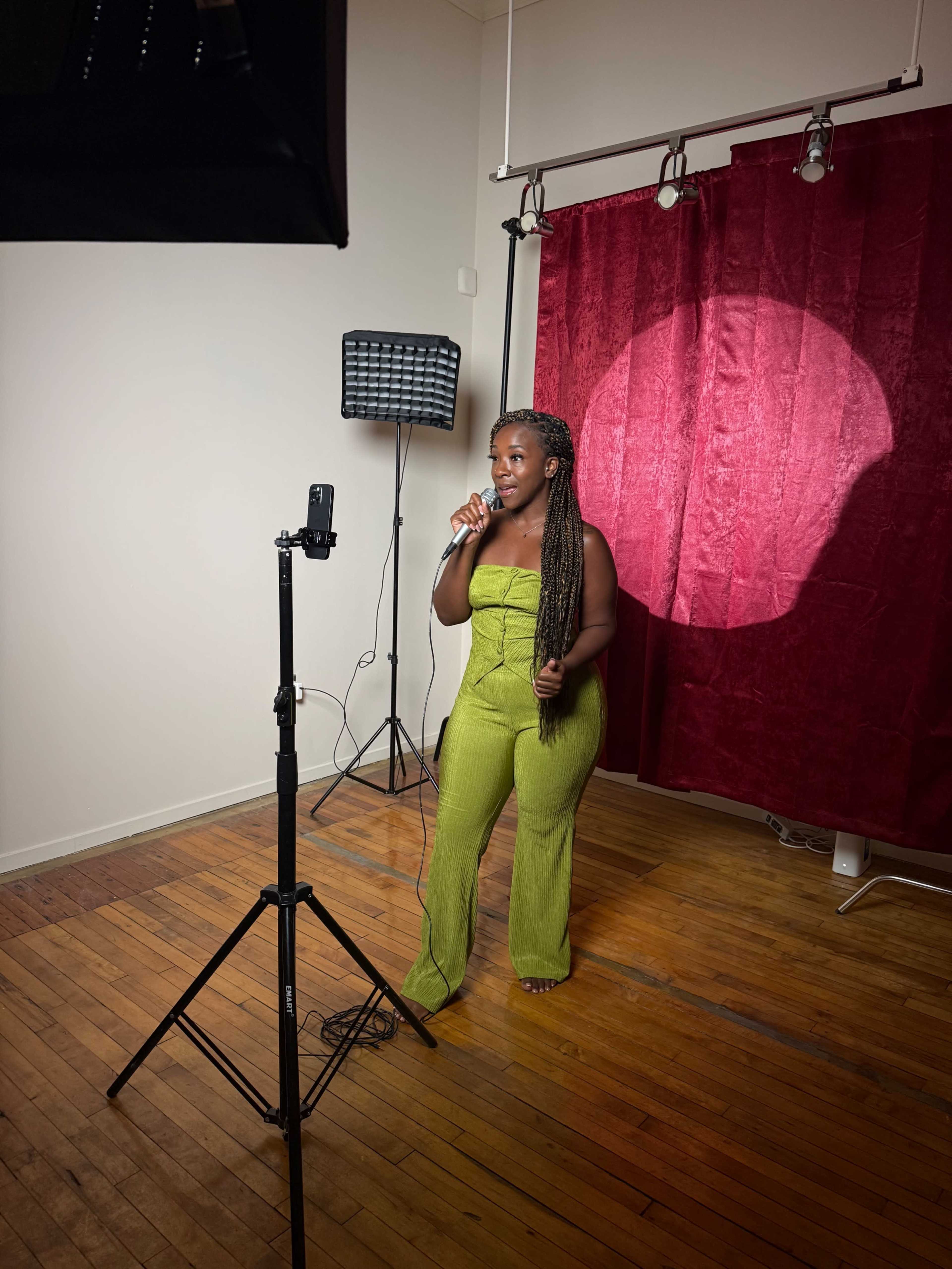 A woman in a green strapless jumpsuit stands holding a microphone in front of a red curtain backdrop, with studio lighting equipment nearby.