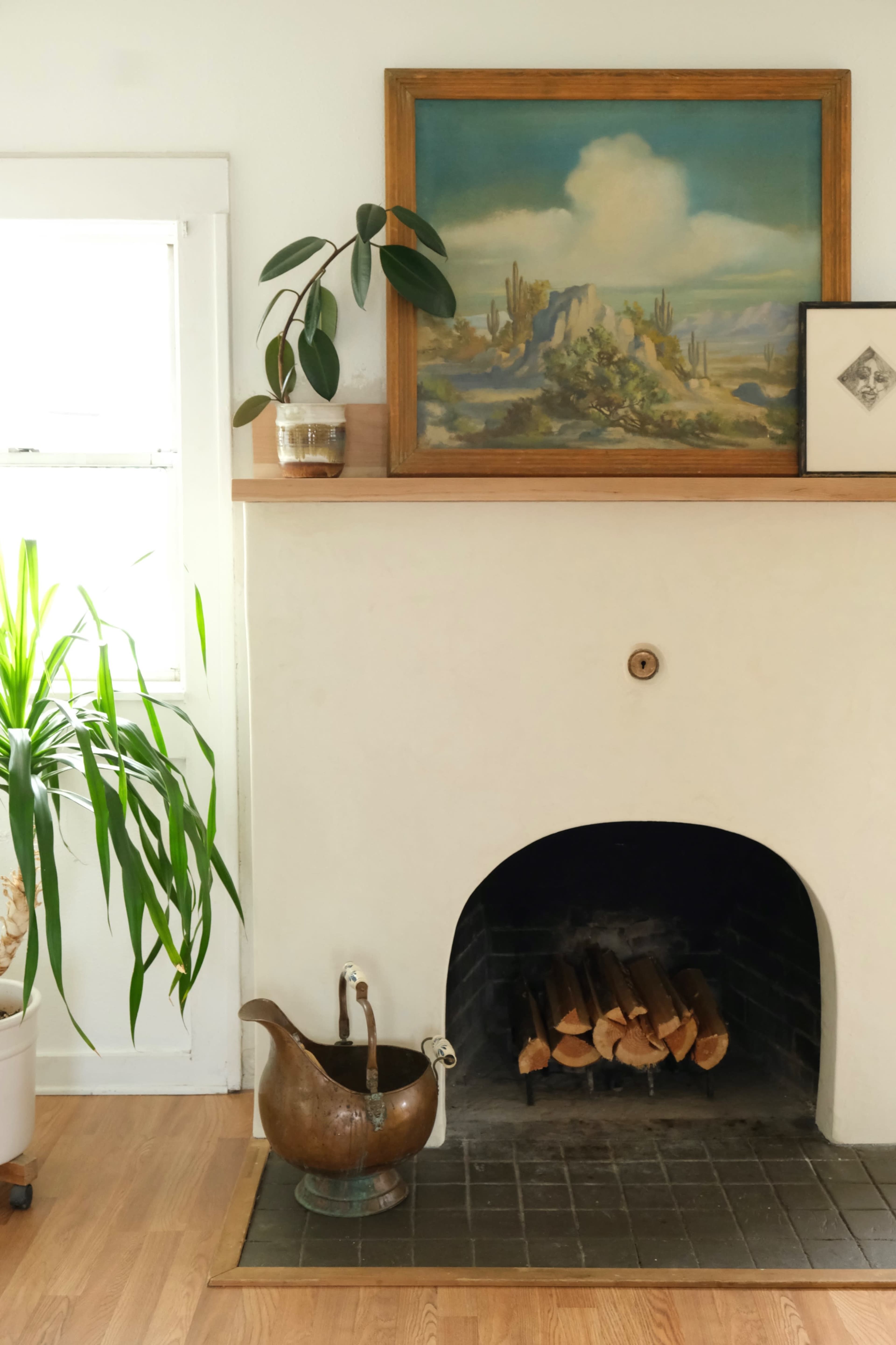 A living room corner featuring a fireplace with stacked logs, a brass pitcher, a framed painting above the mantle, and a potted plant nearby.
