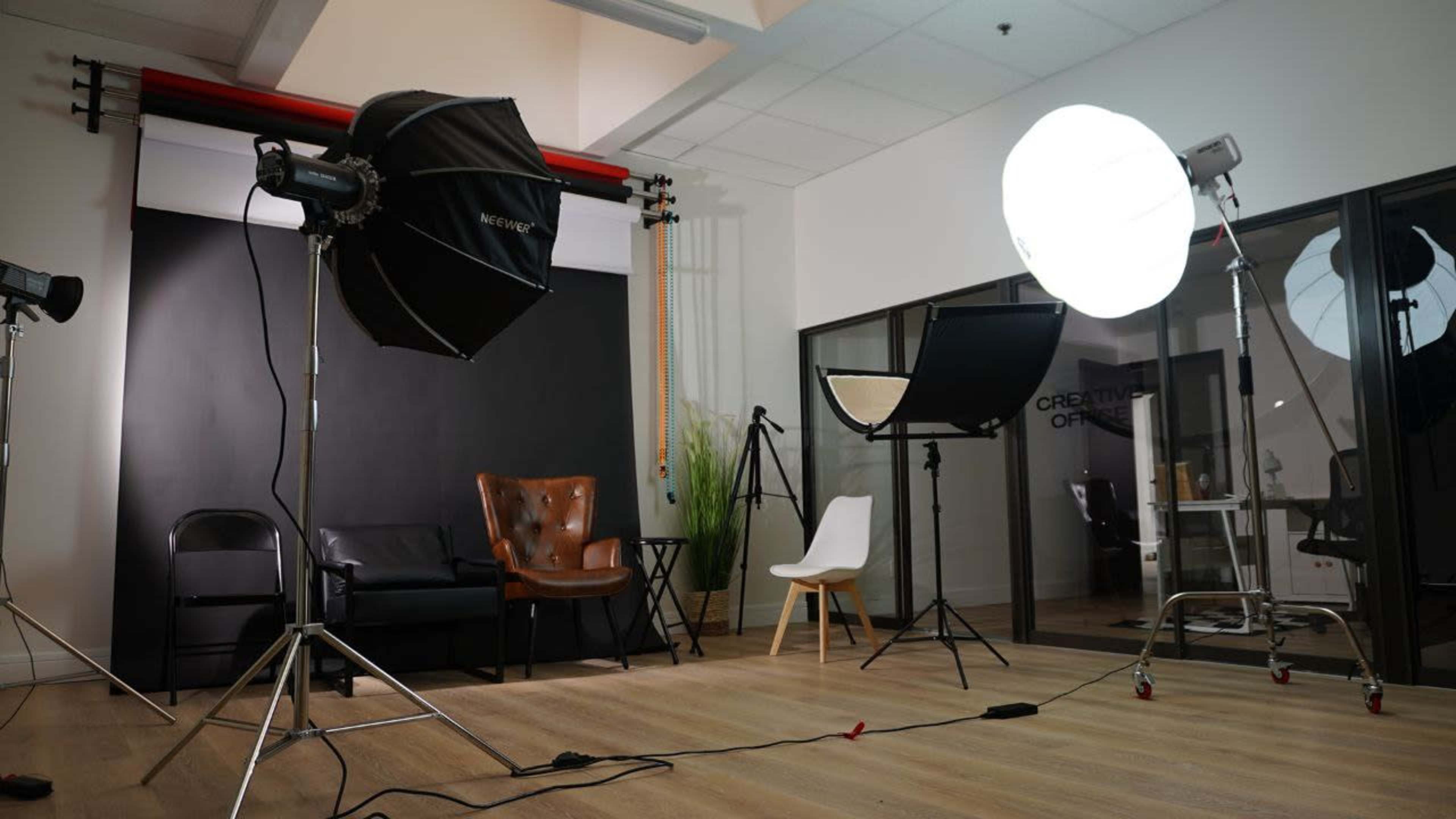 The image shows a photography studio with lighting equipment, a brown leather chair, a white chair, and a black backdrop.