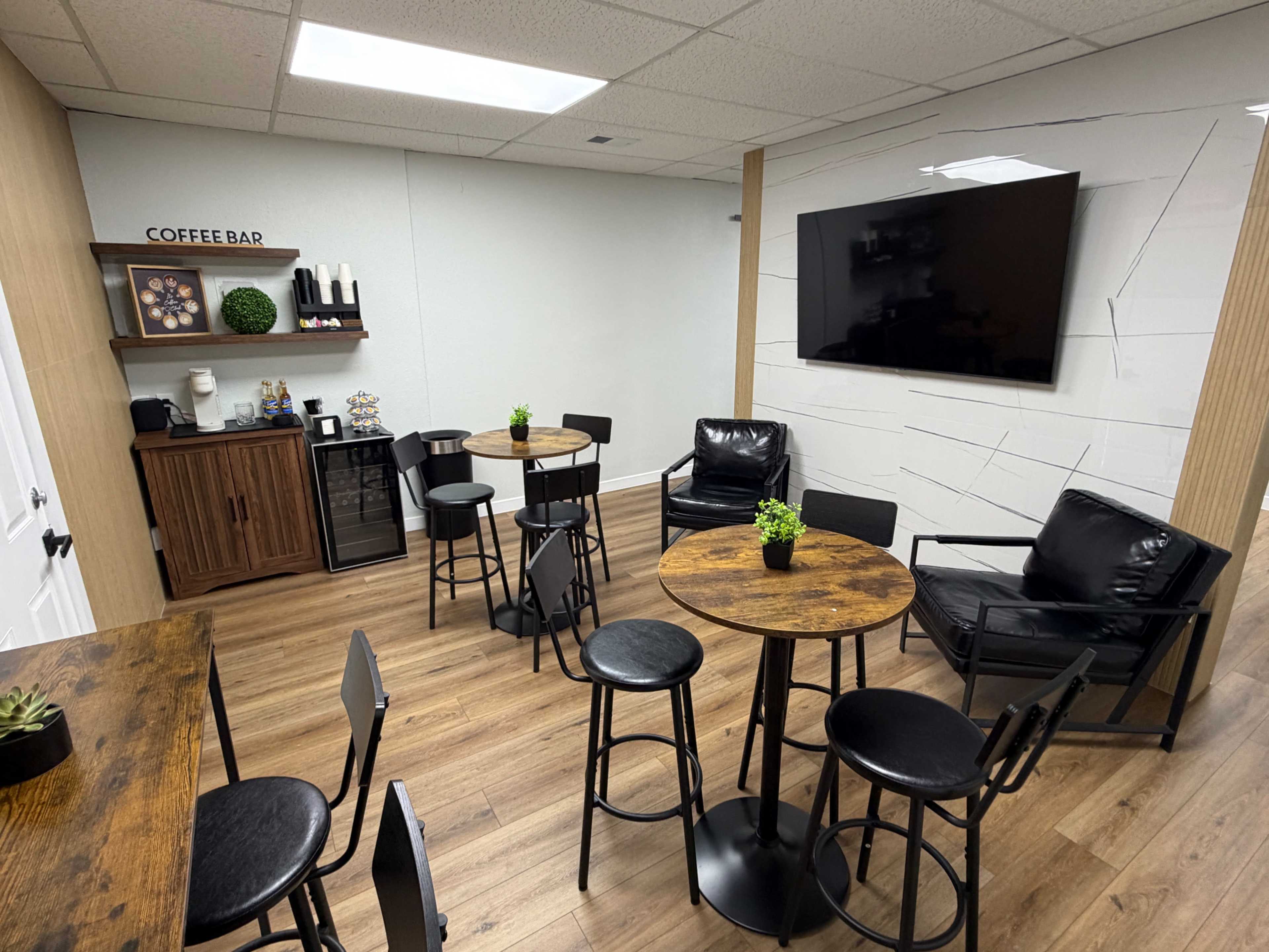 The image shows a lounge area with several round wooden tables, black chairs, and a coffee bar against one wall.