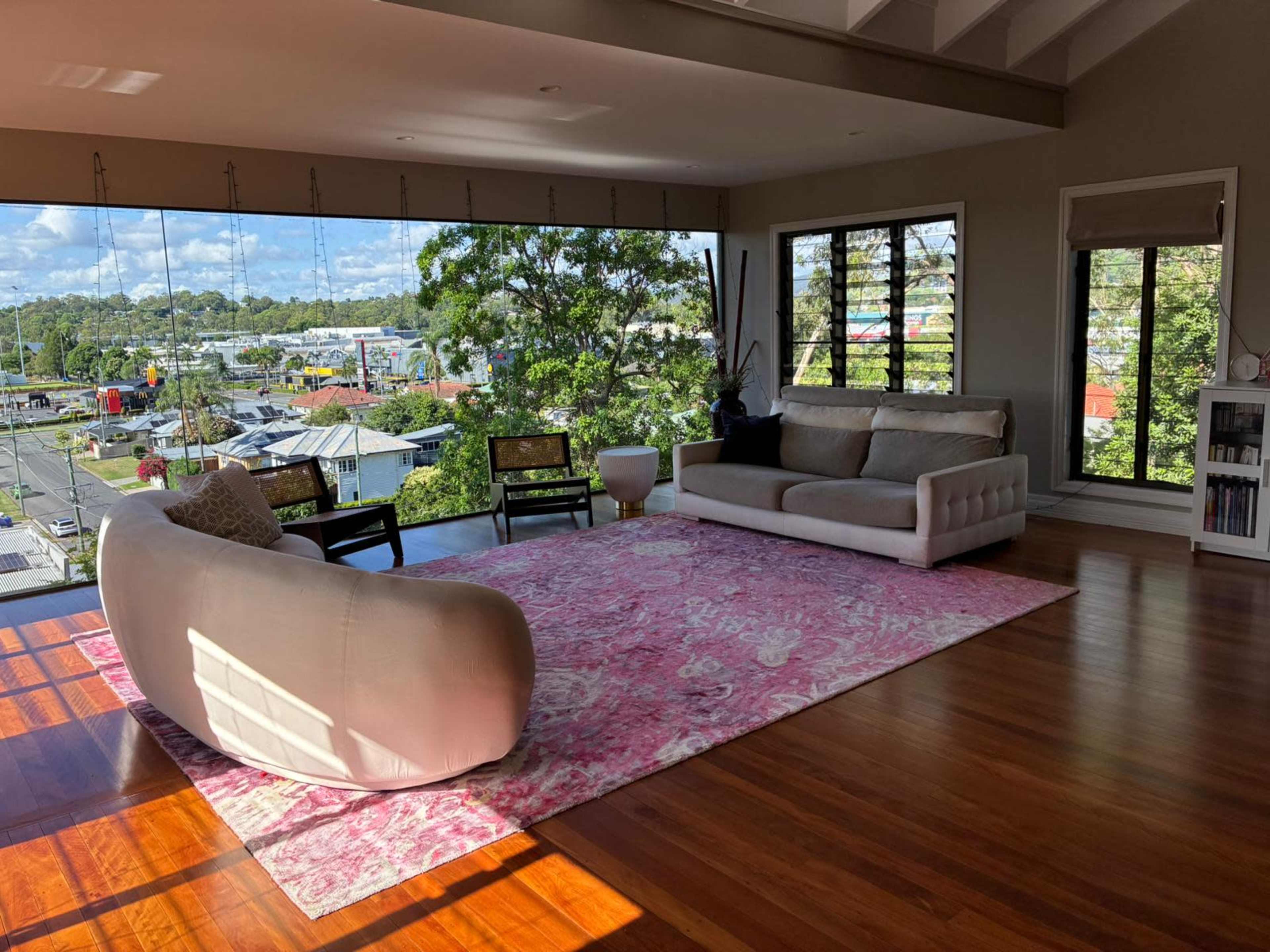 The image shows a modern living room with large windows, a pink area rug, a white sofa, and a view of the outside landscape.