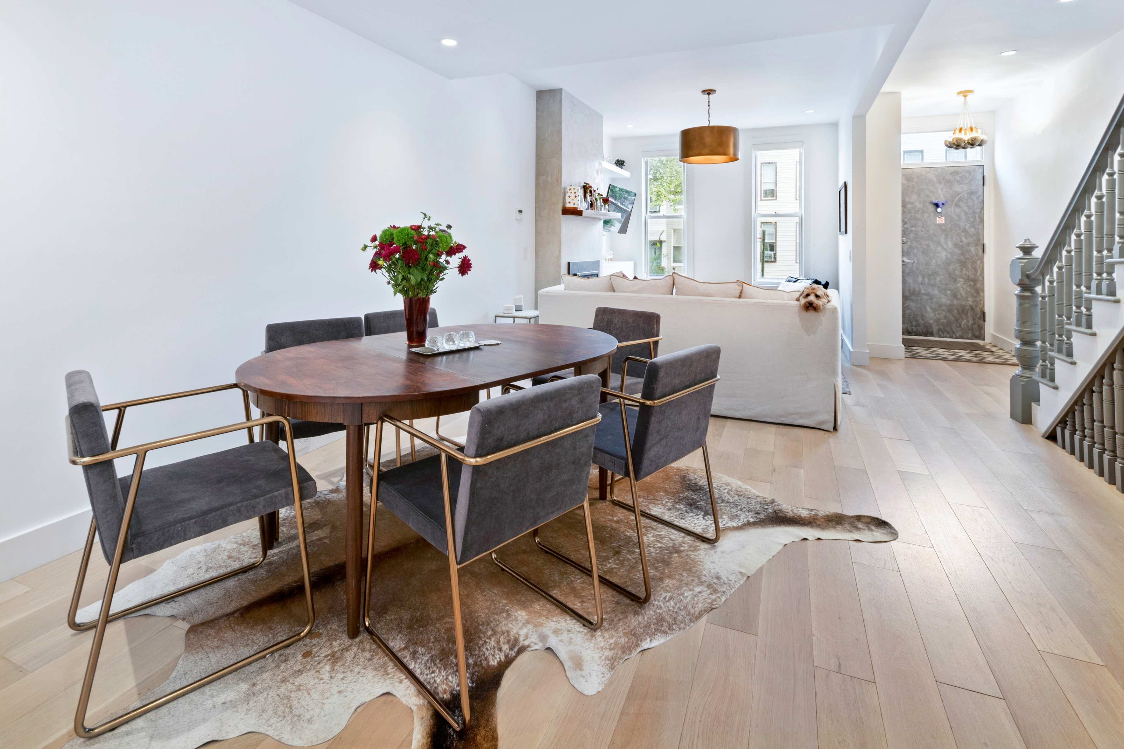 A modern dining area features a round wooden table surrounded by four metal and upholstered chairs, with a decorative rug beneath and a cozy living space visible in the background.