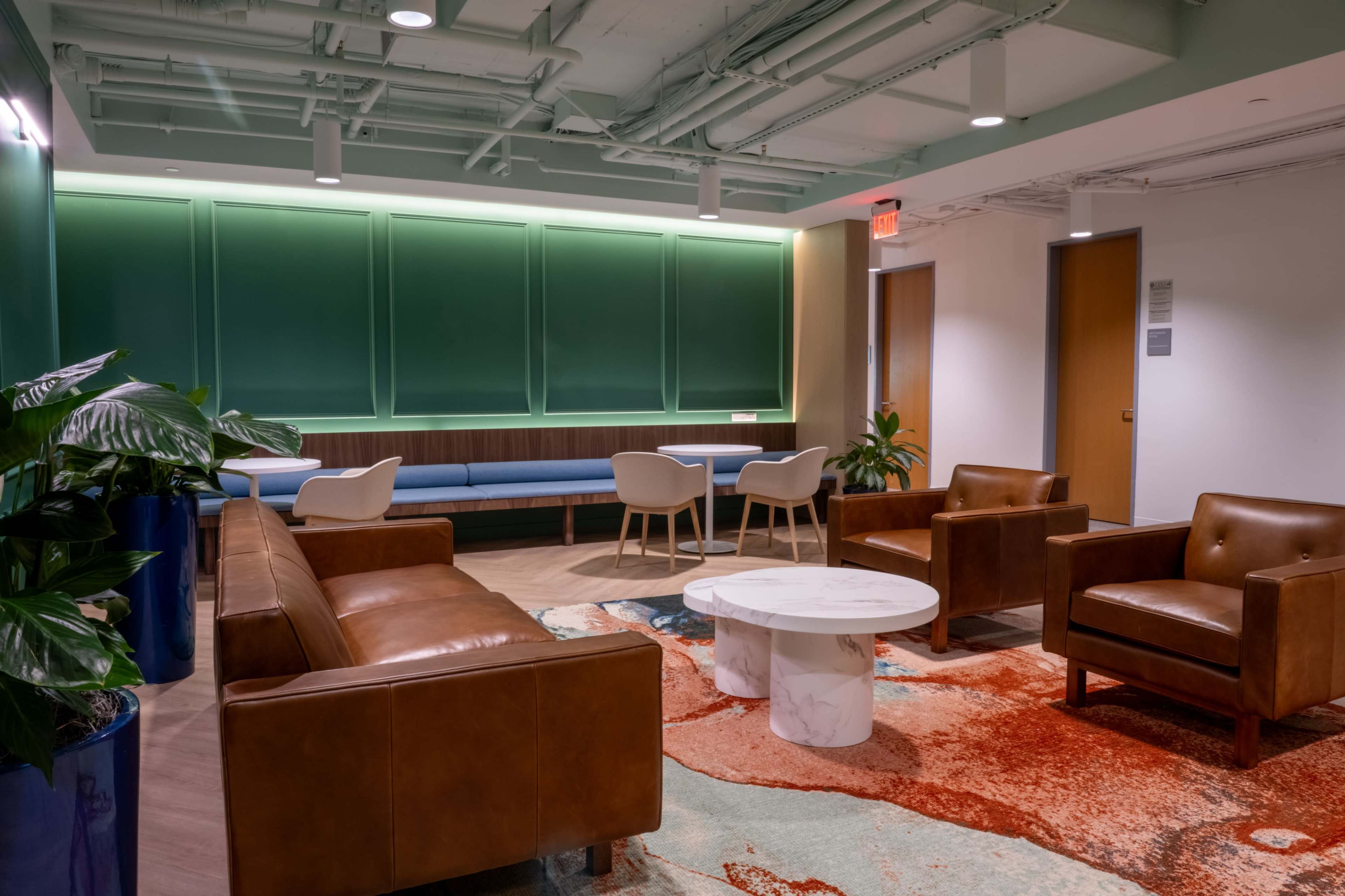 A modern lounge area featuring brown leather chairs, a round marble coffee table, a blue sofa, and green plants, all set against a backdrop of painted green walls.