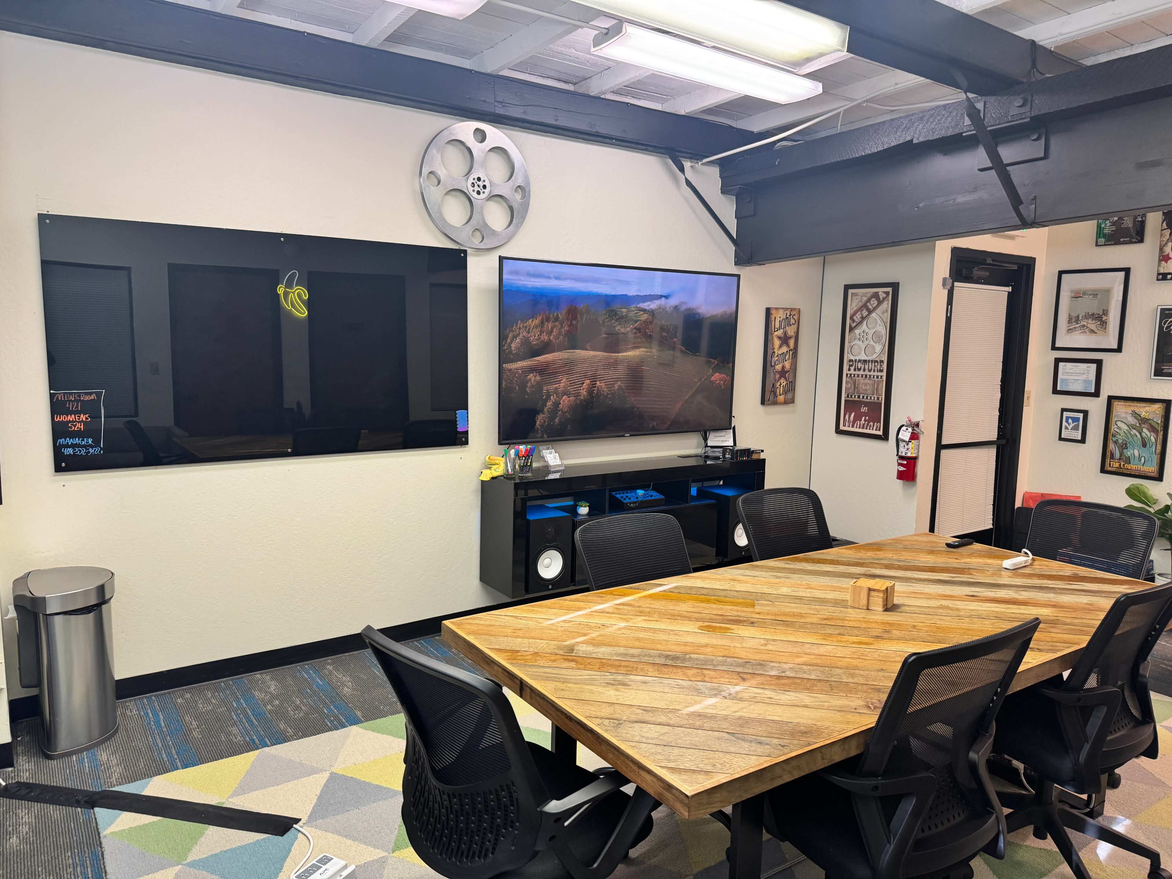 The image shows a modern conference room with a large wooden table, black chairs, a wall-mounted screen, and a projection screen on opposite walls.
