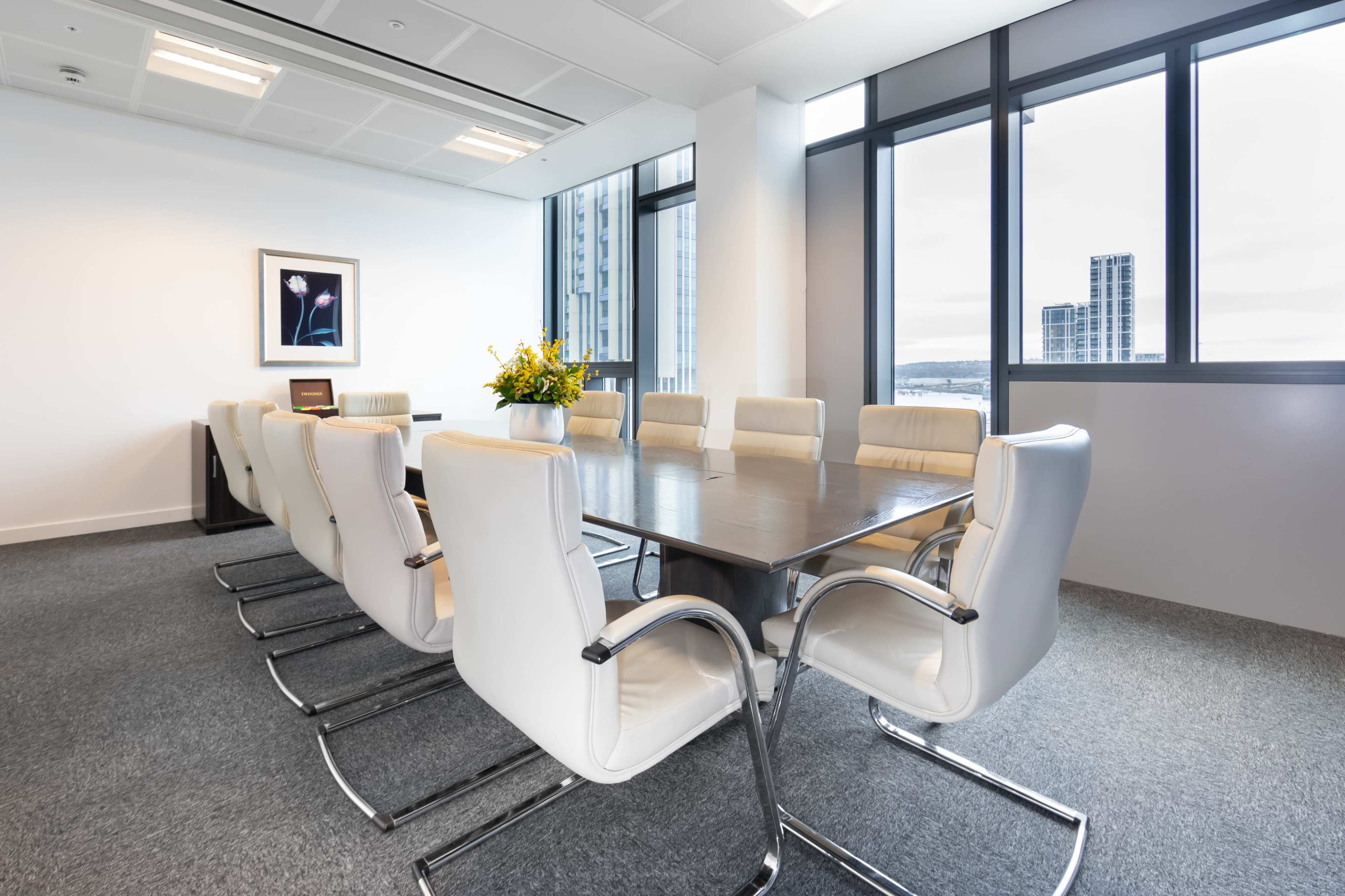 A modern conference room features a long wooden table surrounded by twelve white chairs, with large windows offering a view of the city.