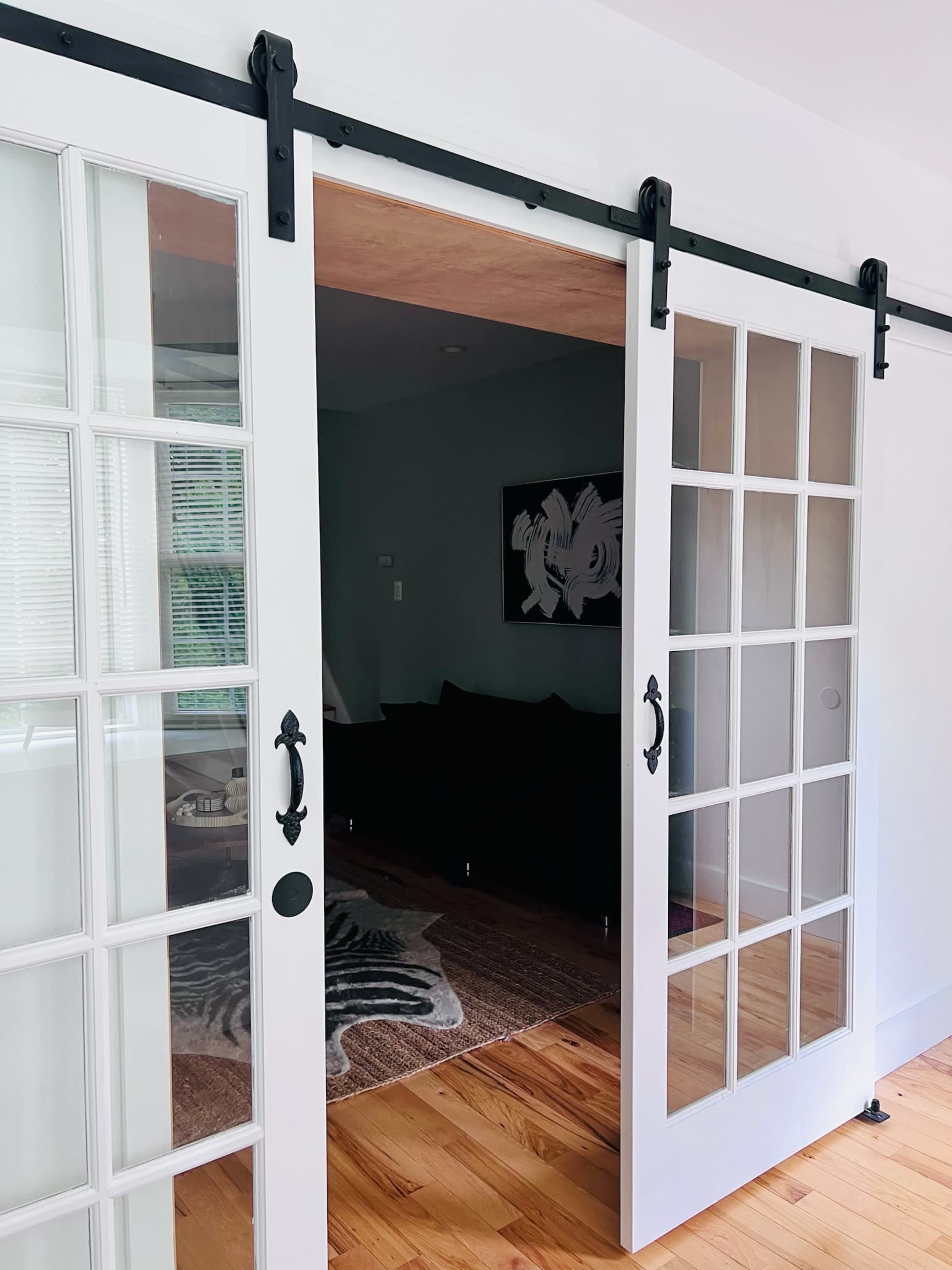 A set of white double barn doors with glass panels, mounted on a black sliding track, leading to a furnished room with a dark couch and a patterned rug.
